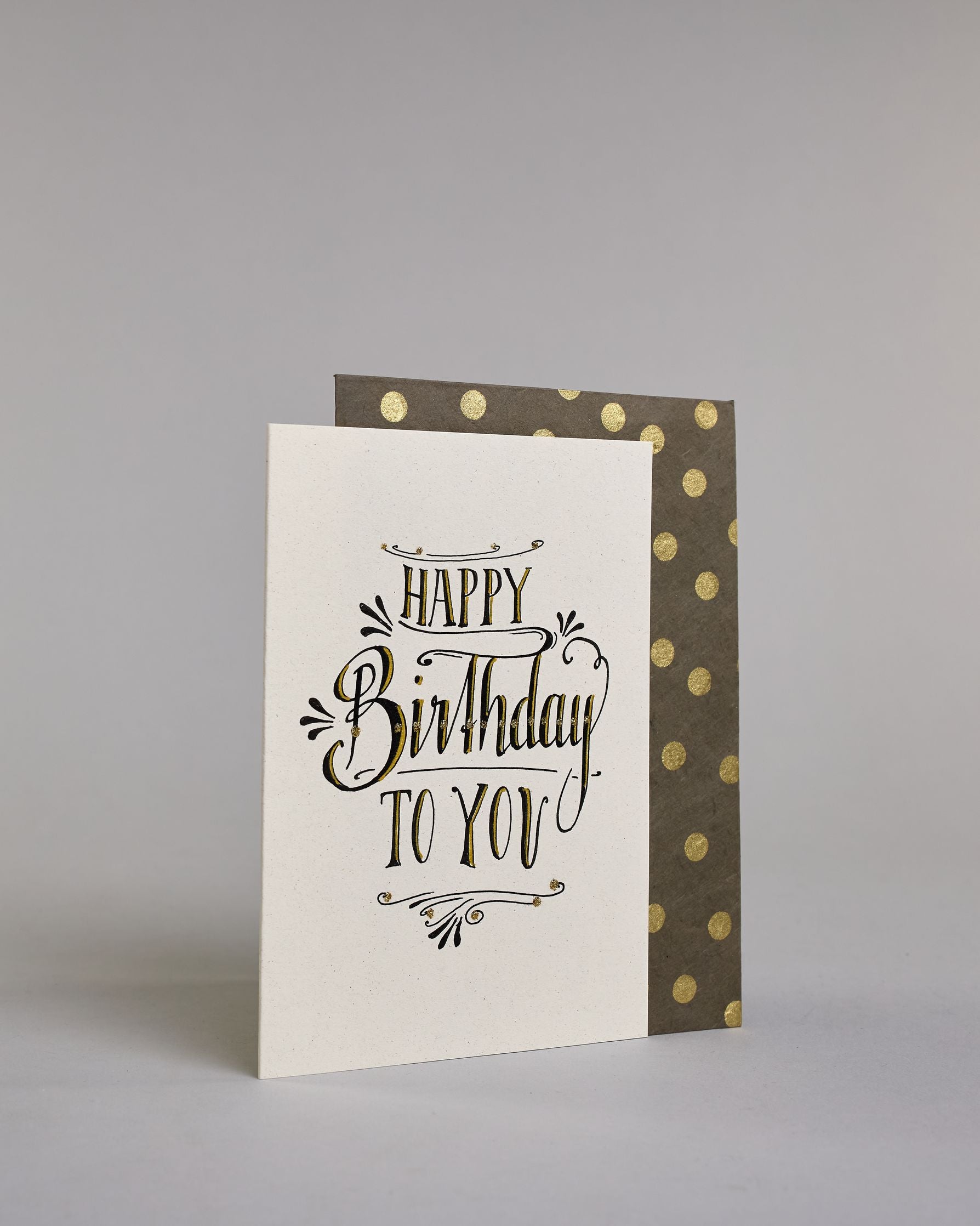 Glückwunschkarte in Englisch zum Geburtstag mit schwarzer Kalligrafieaufschrift "Happy Birthday to you". Der Text ist in drei Zeilen aufgeteilt, welche wiefolge gegliedert sind: "HAPPY", darunter "Birthday", darunter "TO YOU". Die Worte sind mit schwungvollen Linien und Verzierungen umgeben und alles ist mit Gold akzentuiert. Alles ist auf hellem Hintergrund. Dazu ein grün-goldenes handgeschöpftes Kuvert. #3