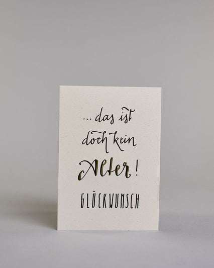 Glückwunschkarte zum Geburtstag mit schwarzer Kalligrafieaufschrift "...das ist doch kein Alter! Glückwunsch". Der Text ist auf vier Zeilen aufgeteilt, welche wiefolge gegliedert sind: "...das ist" in dünner Schreibschrift, darunter "doch kein" in dünner Schreibschrift, darunter "Alter!" in breiterer Schreibschrift und mit Goldakzenten, darunter "GLÜCKWUNSCH" in dünner gerader Kalligrafieschrift. Alles ist auf hellem Hintergrund. Dazu ein grün-goldenes handgeschöpftes Kuvert. #1