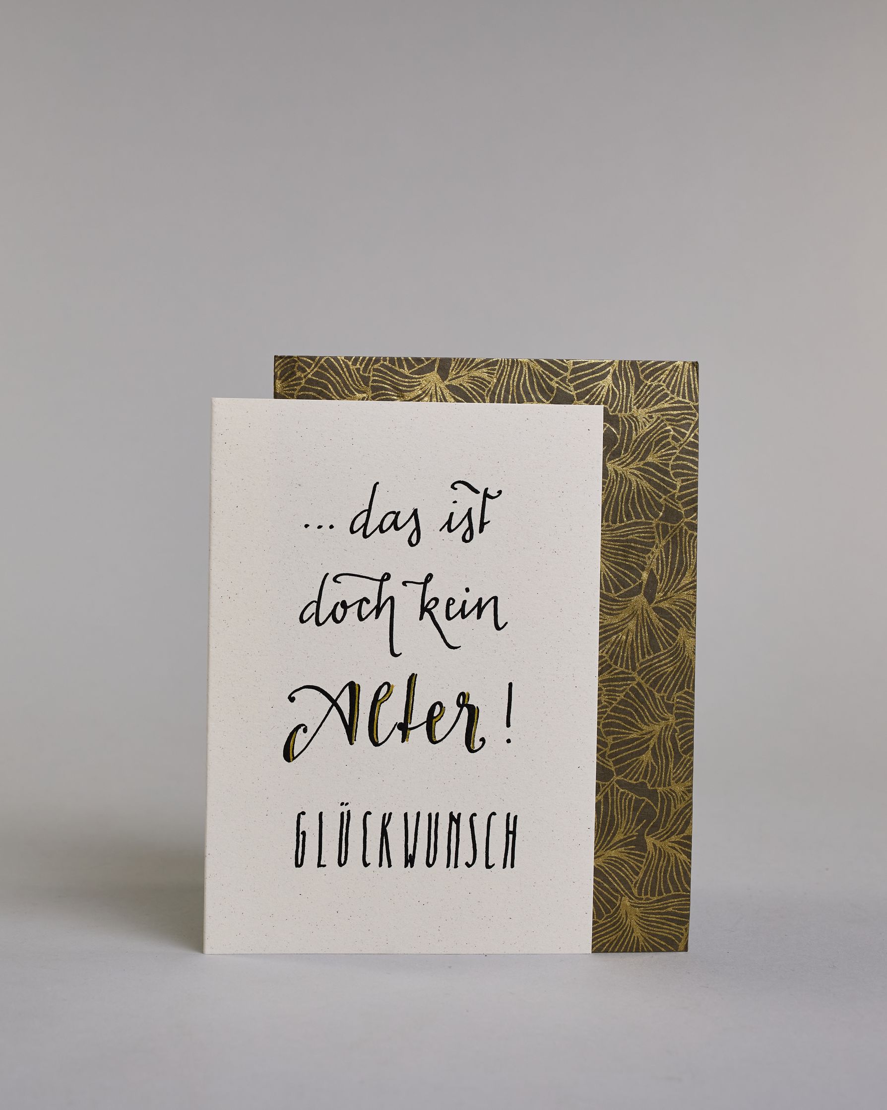 Glückwunschkarte zum Geburtstag mit schwarzer Kalligrafieaufschrift "...das ist doch kein Alter! Glückwunsch". Der Text ist auf vier Zeilen aufgeteilt, welche wiefolge gegliedert sind: "...das ist" in dünner Schreibschrift, darunter "doch kein" in dünner Schreibschrift, darunter "Alter!" in breiterer Schreibschrift und mit Goldakzenten, darunter "GLÜCKWUNSCH" in dünner gerader Kalligrafieschrift. Alles ist auf hellem Hintergrund. Dazu ein grün-goldenes handgeschöpftes Kuvert. #2