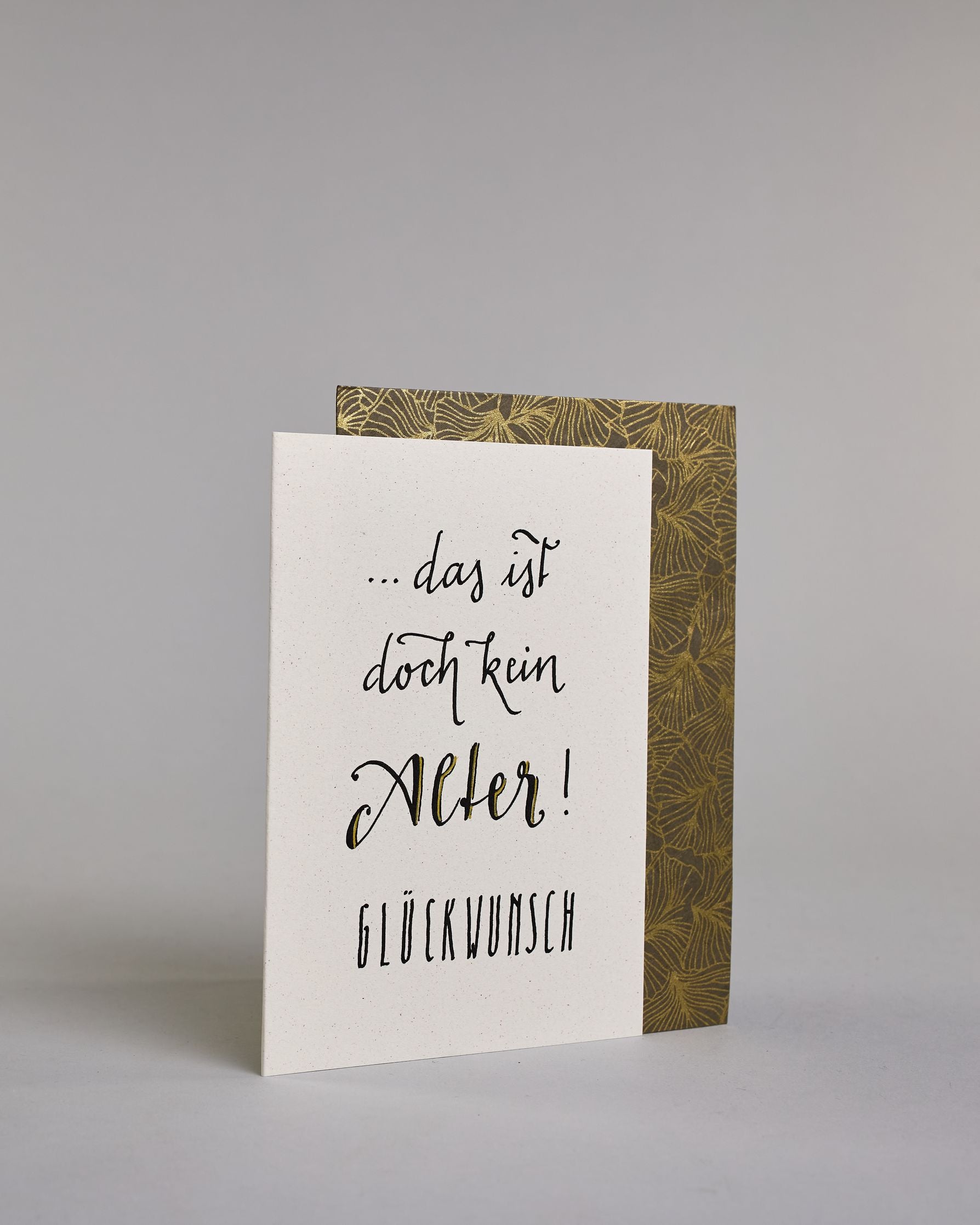 Glückwunschkarte zum Geburtstag mit schwarzer Kalligrafieaufschrift "...das ist doch kein Alter! Glückwunsch". Der Text ist auf vier Zeilen aufgeteilt, welche wiefolge gegliedert sind: "...das ist" in dünner Schreibschrift, darunter "doch kein" in dünner Schreibschrift, darunter "Alter!" in breiterer Schreibschrift und mit Goldakzenten, darunter "GLÜCKWUNSCH" in dünner gerader Kalligrafieschrift. Alles ist auf hellem Hintergrund. Dazu ein grün-goldenes handgeschöpftes Kuvert. #3