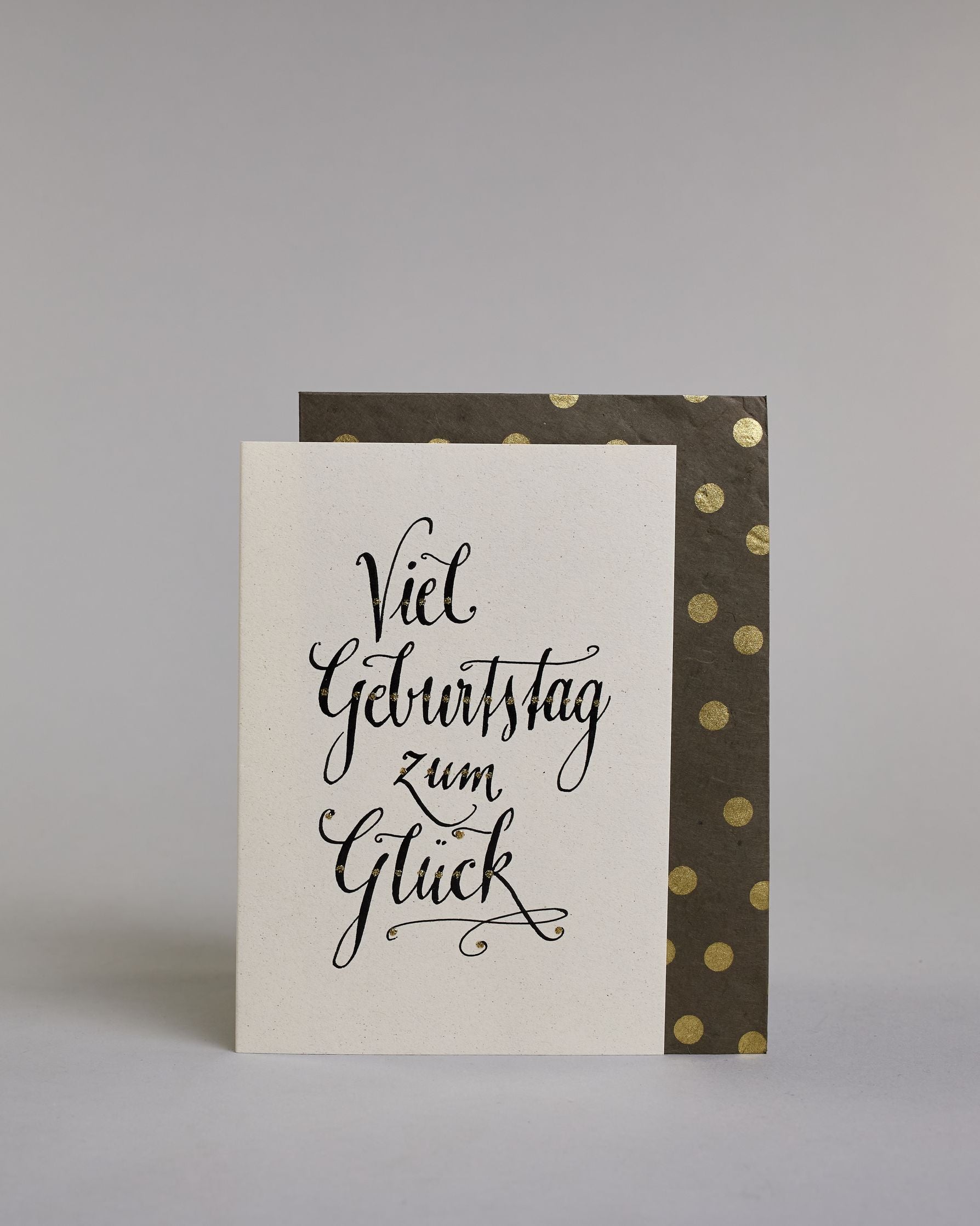 Glückwunschkarte zum Geburtstag mit schwarzer Kalligrafieaufschrift "Viel Geburtstag zum Glück". Das Wortspiel ist auf vier Zeilen aufgeteilt, welche jeweils immer ein Wort beinhalten. Die Schrift ist eine große geschwungene Schreibschrift mit Goldakzenten auf hellem Hintergrund. Dazu ein grün-goldenes handgeschöpftes Kuvert. #2