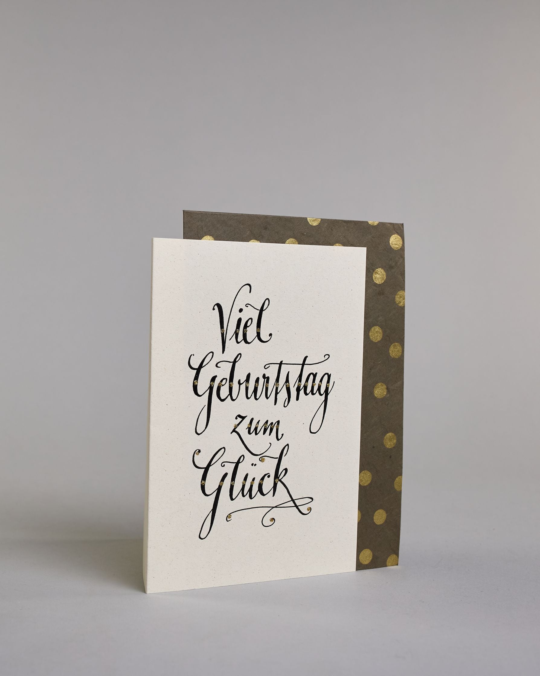 Glückwunschkarte zum Geburtstag mit schwarzer Kalligrafieaufschrift "Viel Geburtstag zum Glück". Das Wortspiel ist auf vier Zeilen aufgeteilt, welche jeweils immer ein Wort beinhalten. Die Schrift ist eine große geschwungene Schreibschrift mit Goldakzenten auf hellem Hintergrund. Dazu ein grün-goldenes handgeschöpftes Kuvert. #3
