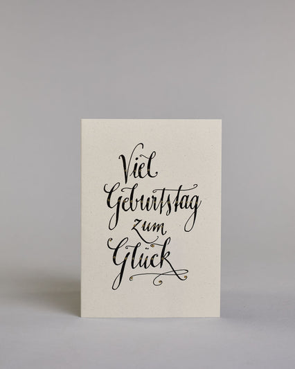 Glückwunschkarte zum Geburtstag mit schwarzer Kalligrafieaufschrift "Viel Geburtstag zum Glück". Das Wortspiel ist auf vier Zeilen aufgeteilt, welche jeweils immer ein Wort beinhalten. Die Schrift ist eine große geschwungene Schreibschrift mit Goldakzenten auf hellem Hintergrund. Dazu ein grün-goldenes handgeschöpftes Kuvert. #1
