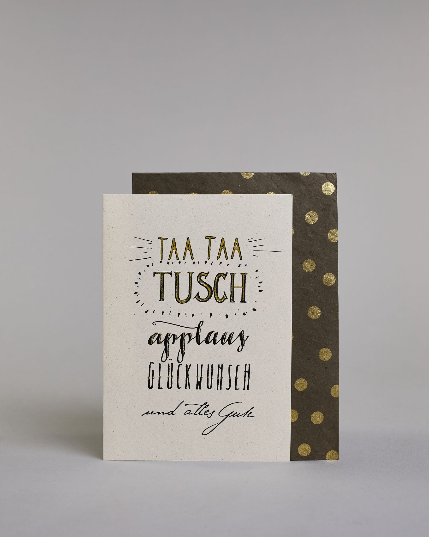 Glückwunschkarte zum Geburtstag mit schwarzer Kalligrafieaufschrift "Taa taa tusch applaus Glückwunsch und alles Gute". Die Worte sind auf fünf Zeilen aufgeteilt, welche wiefolgt aufgeteilt sind: "TAA TAA", darunter "TUSCH", beide Zeilen sind in Blockbuchstaben und die Buchstaben sind mit Gold gefüllt, rundherum sind dekorative Strahlen, darunter in geschwungener Schreibschrift "applaus", darunter "GLÜCKWUNSCH" in dünner, gerader Kalligrafieschrift, darunter "und alles Gute" in kleiner, dünner Schreibschri…