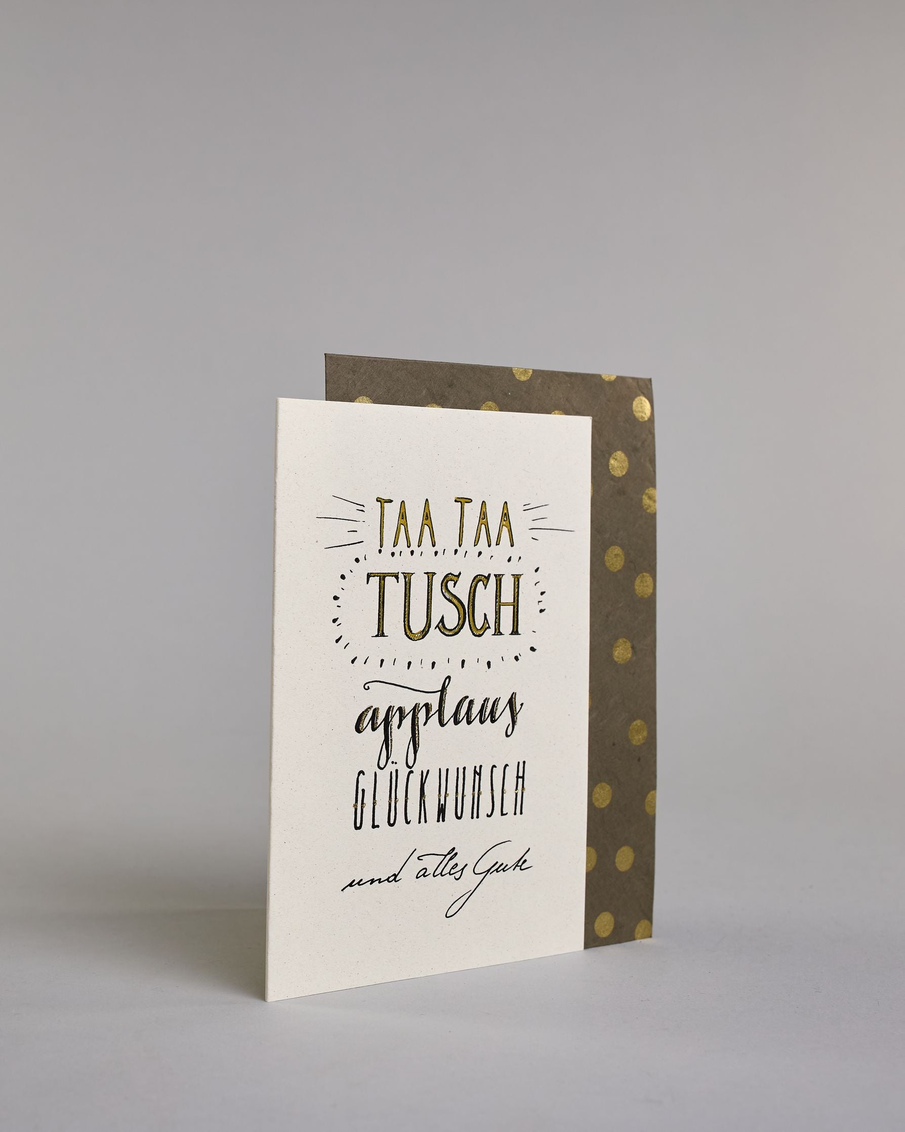 Glückwunschkarte zum Geburtstag mit schwarzer Kalligrafieaufschrift "Taa taa tusch applaus Glückwunsch und alles Gute". Die Worte sind auf fünf Zeilen aufgeteilt, welche wiefolgt aufgeteilt sind: "TAA TAA", darunter "TUSCH", beide Zeilen sind in Blockbuchstaben und die Buchstaben sind mit Gold gefüllt, rundherum sind dekorative Strahlen, darunter in geschwungener Schreibschrift "applaus", darunter "GLÜCKWUNSCH" in dünner, gerader Kalligrafieschrift, darunter "und alles Gute" in kleiner, dünner Schreibschri…