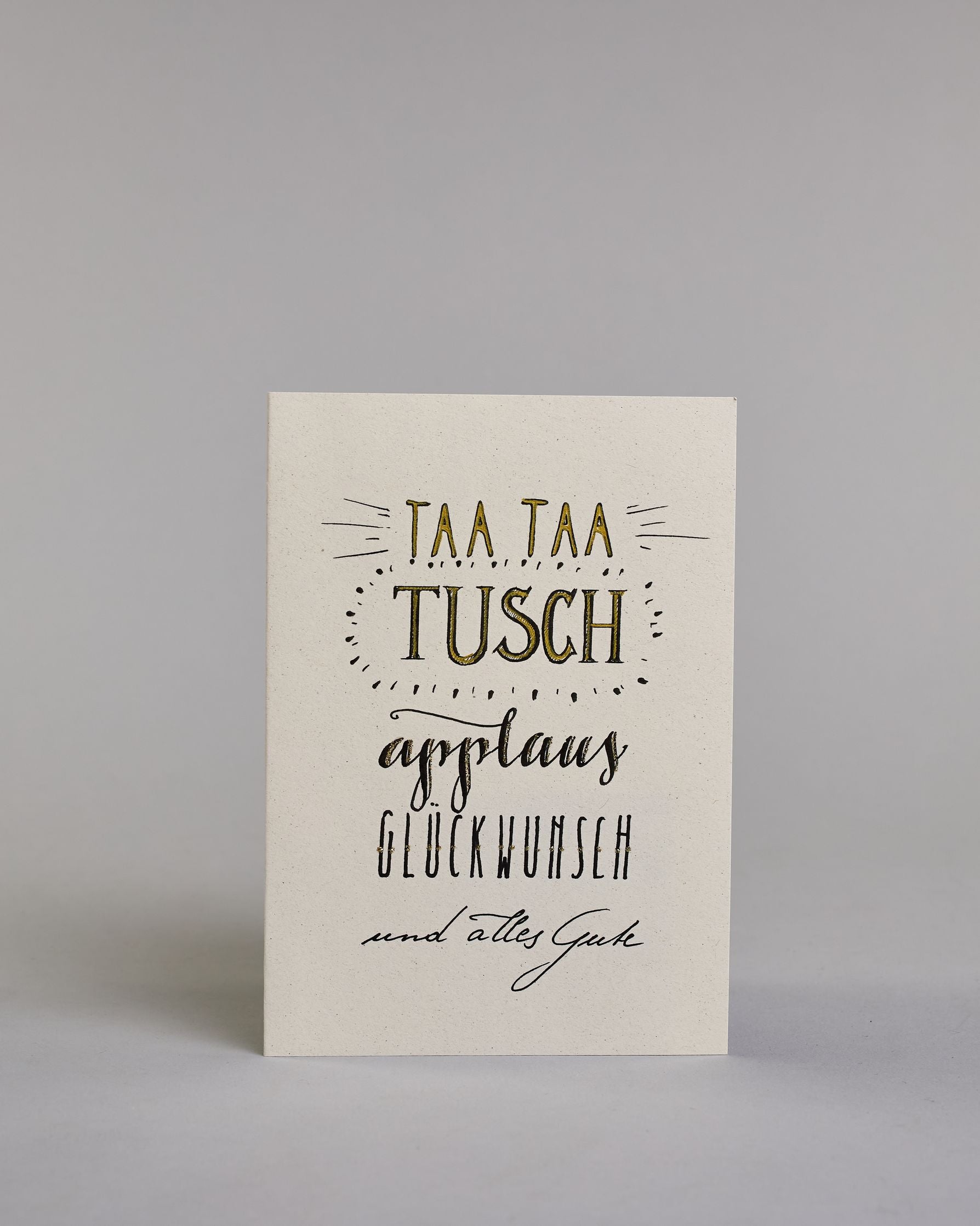 Glückwunschkarte zum Geburtstag mit schwarzer Kalligrafieaufschrift "Taa taa tusch applaus Glückwunsch und alles Gute". Die Worte sind auf fünf Zeilen aufgeteilt, welche wiefolgt aufgeteilt sind: "TAA TAA", darunter "TUSCH", beide Zeilen sind in Blockbuchstaben und die Buchstaben sind mit Gold gefüllt, rundherum sind dekorative Strahlen, darunter in geschwungener Schreibschrift "applaus", darunter "GLÜCKWUNSCH" in dünner, gerader Kalligrafieschrift, darunter "und alles Gute" in kleiner, dünner Schreibschri…