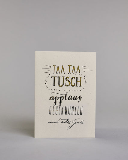 Glückwunschkarte zum Geburtstag mit schwarzer Kalligrafieaufschrift "Taa taa tusch applaus Glückwunsch und alles Gute". Die Worte sind auf fünf Zeilen aufgeteilt, welche wiefolgt aufgeteilt sind: "TAA TAA", darunter "TUSCH", beide Zeilen sind in Blockbuchstaben und die Buchstaben sind mit Gold gefüllt, rundherum sind dekorative Strahlen, darunter in geschwungener Schreibschrift "applaus", darunter "GLÜCKWUNSCH" in dünner, gerader Kalligrafieschrift, darunter "und alles Gute" in kleiner, dünner Schreibschri…