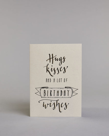 Glückwunschkarte auf Englisch zum Geburtstag mit der Aufschrift "Hugs kisses and a lot of BIRTHDAY wishes" in schwarzer Schrift auf hellem Hintergrund. Die ersten zwei Zeilen, welche jeweils das Wort "Hugs" und "kisses" beinhaltet sind in der gleichen etwas dickeren schwarzen Schreibschrift gestaltet, darunter "AND A LOT OF" in einer dünnen geraden Schrift, darunter das Wort "BIRTHDAY" ist in dünn geschriebenen Großbuchstaben in einem gezeichneten Banner hervorgehoben, darunter dann noch in der gleichen Sc…