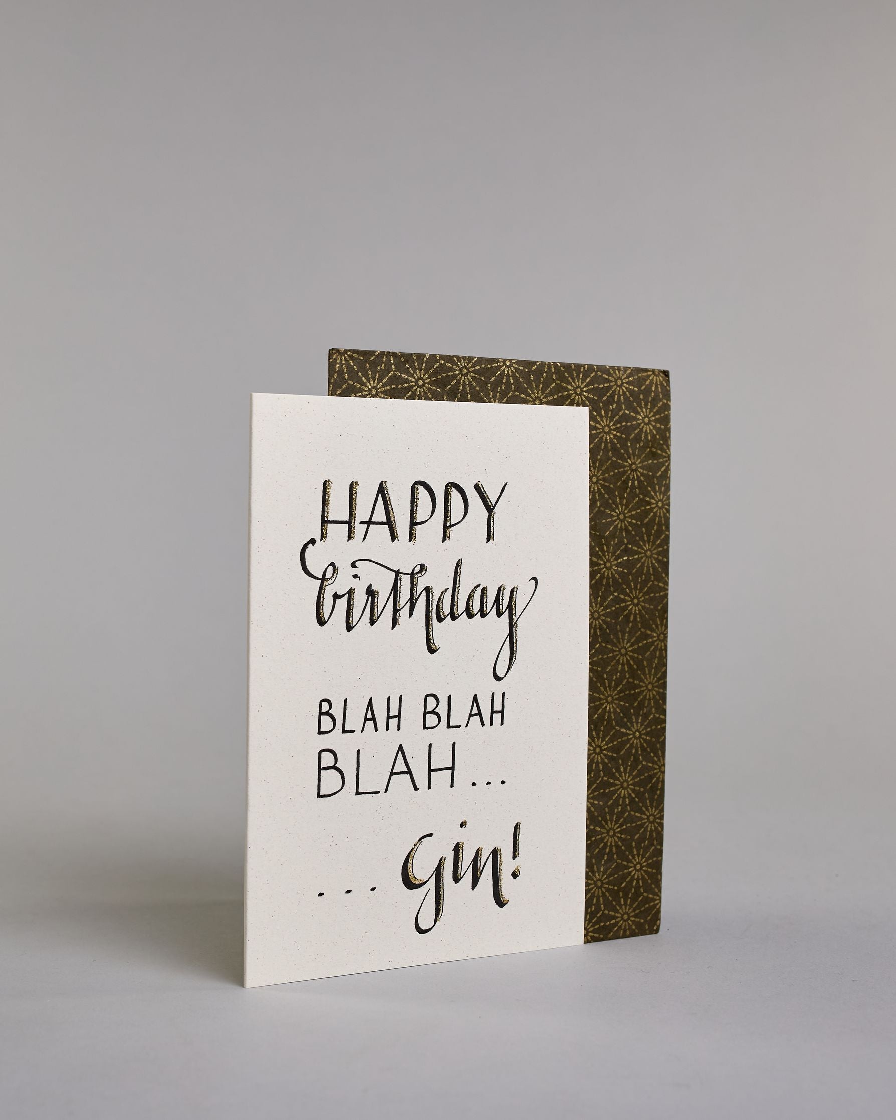 Glückwunschkarte auf Englisch zum Geburtstag mit der Aufschrift "Happy birthday blah blah blah ...gin!" in schwarzer, goldakzentuierter Schrift auf hellem Hintergrund. Die Zeilen sind wiefolgt aufgeteilt: "HAPPY", darunter "birthday" beides in eher dickerer Schrift, darunter "BLAH BLAH", darunter "BLAH..." beide Zeilen in dünner gerader Kalligrafieschrift,  darunter "...Gin!" wie in geschwungener dickeren Schrift. Dazu ein grün-goldenes handgeschöpftes Kuvert. #3