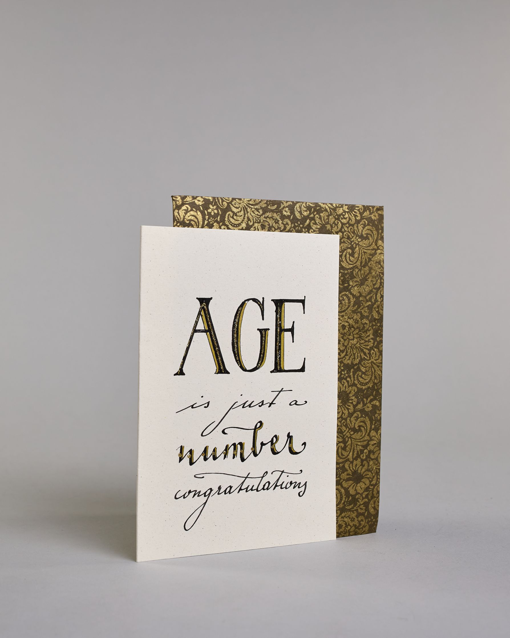 Glückwunschkarte auf Englisch zum Geburtstag mit der Aufschrift "Age ist just a number congratulations" in schwarzer, goldakzentuierter Schrift auf hellem Hintergrund. Die Zeilen sind wiefolgt aufgeteilt: "AGE" in großen Blockbuchstaben, darunter "is just a" in dünner geschwungener Schrift, darunter "number" in dicker geschwungener Schrift, darunter "congratulations" wieder in dünner geschwungener Kalligrafieschrift. Dazu ein grün-goldenes handgeschöpftes Kuvert. #3