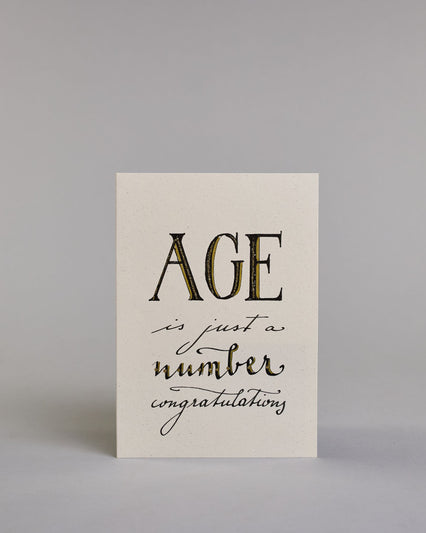 Glückwunschkarte auf Englisch zum Geburtstag mit der Aufschrift "Age ist just a number congratulations" in schwarzer, goldakzentuierter Schrift auf hellem Hintergrund. Die Zeilen sind wiefolgt aufgeteilt: "AGE" in großen Blockbuchstaben, darunter "is just a" in dünner geschwungener Schrift, darunter "number" in dicker geschwungener Schrift, darunter "congratulations" wieder in dünner geschwungener Kalligrafieschrift. Dazu ein grün-goldenes handgeschöpftes Kuvert. #1