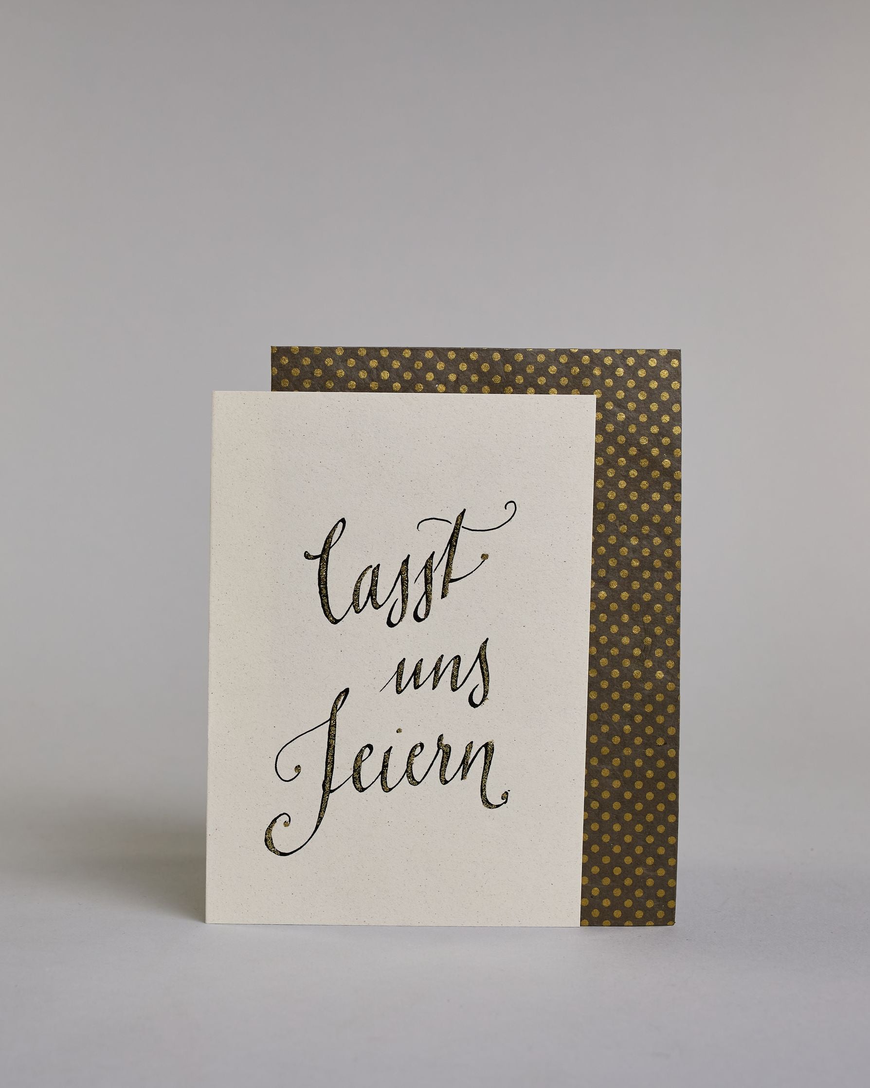 Glückwunschkarte zum Geburtstag mit schwarzer Kalligrafieaufschrift "Lasst uns feiern". Der Text ist auf drei Zeilen aufgeteilt, welche jeweils immer ein Wort beinhalten. Die Schrift ist eine große geschwungene Schreibschrift mit Goldakzenten auf hellem Hintergrund. Dazu ein grün-goldenes handgeschöpftes Kuvert. #2