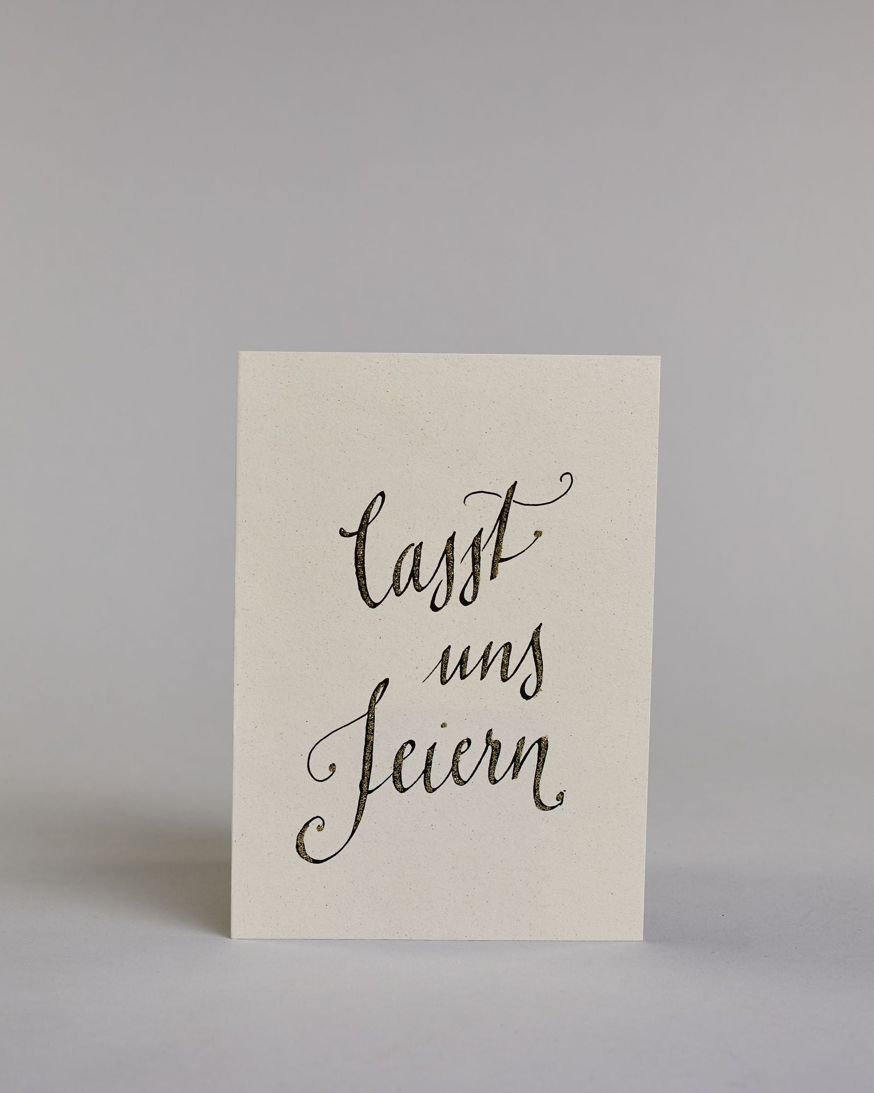 Glückwunschkarte zum Geburtstag mit schwarzer Kalligrafieaufschrift "Lasst uns feiern". Der Text ist auf drei Zeilen aufgeteilt, welche jeweils immer ein Wort beinhalten. Die Schrift ist eine große geschwungene Schreibschrift mit Goldakzenten auf hellem Hintergrund. Dazu ein grün-goldenes handgeschöpftes Kuvert. #1