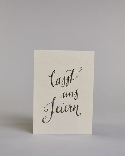Glückwunschkarte zum Geburtstag mit schwarzer Kalligrafieaufschrift "Lasst uns feiern". Der Text ist auf drei Zeilen aufgeteilt, welche jeweils immer ein Wort beinhalten. Die Schrift ist eine große geschwungene Schreibschrift mit Goldakzenten auf hellem Hintergrund. Dazu ein grün-goldenes handgeschöpftes Kuvert. #1