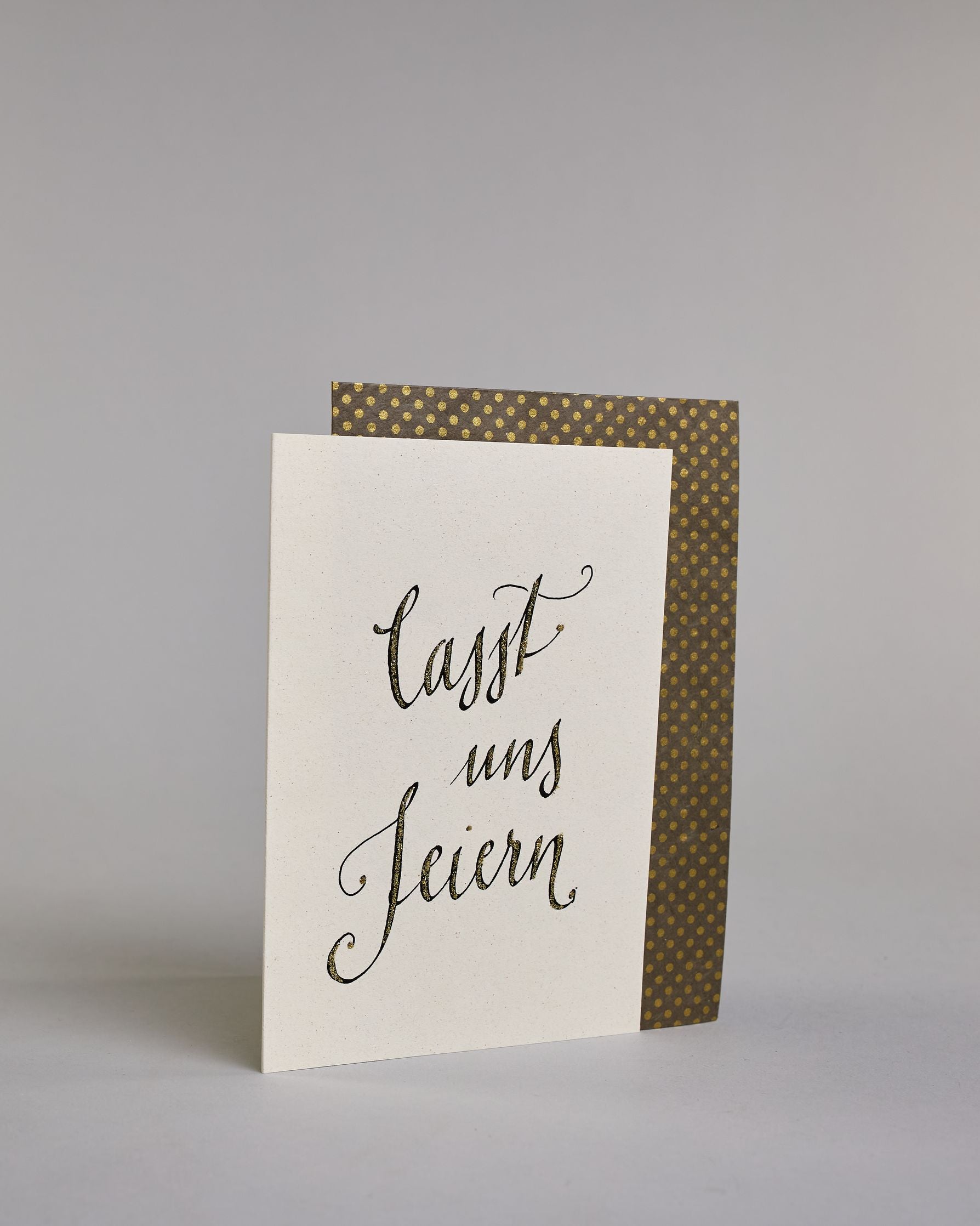 Glückwunschkarte zum Geburtstag mit schwarzer Kalligrafieaufschrift "Lasst uns feiern". Der Text ist auf drei Zeilen aufgeteilt, welche jeweils immer ein Wort beinhalten. Die Schrift ist eine große geschwungene Schreibschrift mit Goldakzenten auf hellem Hintergrund. Dazu ein grün-goldenes handgeschöpftes Kuvert. #3