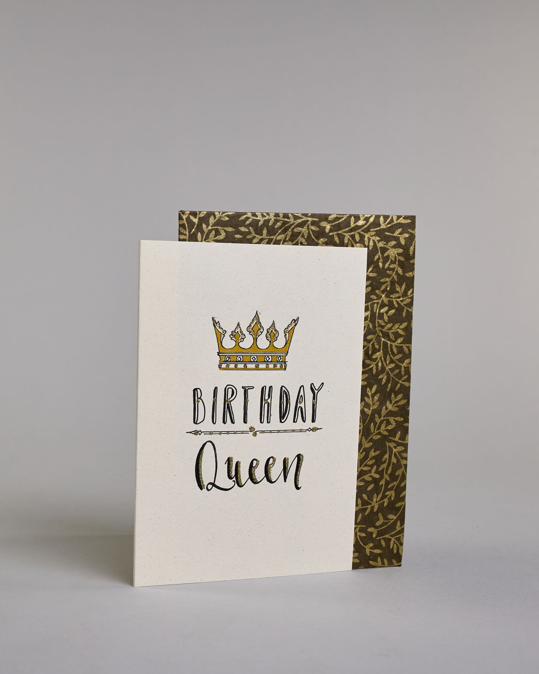 Glückwunschkarte auf Englisch zum Geburtstag mit schwarzer Kalligrafieaufschrift "Birthday Queen", mit einer prunkvollen goldenen und goldglitzernden Krone. Der Text ist übereinander geschrieben mit einer horizontalen goldverzierten Linie dazwischen. Das "Birthday" ist in Blockbuchstaben und das "Queen" in geschwungener Schreibschrift auf hellem Hintergrund. Dazu ein grün-goldenes handgeschöpftes Kuvert. #3