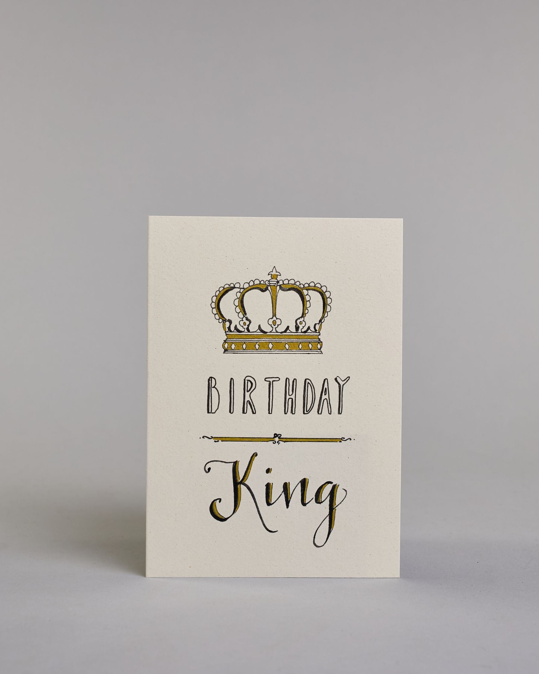 Glückwunschkarte auf Englisch zum Geburtstag mit schwarzer Kalligrafieaufschrift "Birthday King", mit einer prunkvollen goldenen und goldglitzernden Krone. Der Text ist übereinander geschrieben mit einer horizontalen goldverzierten Linie dazwischen. Das "Birthday" ist in Blockbuchstaben und das "King" in geschwungener Schreibschrift auf hellem Hintergrund. Dazu ein grün-goldenes handgeschöpftes Kuvert. #1