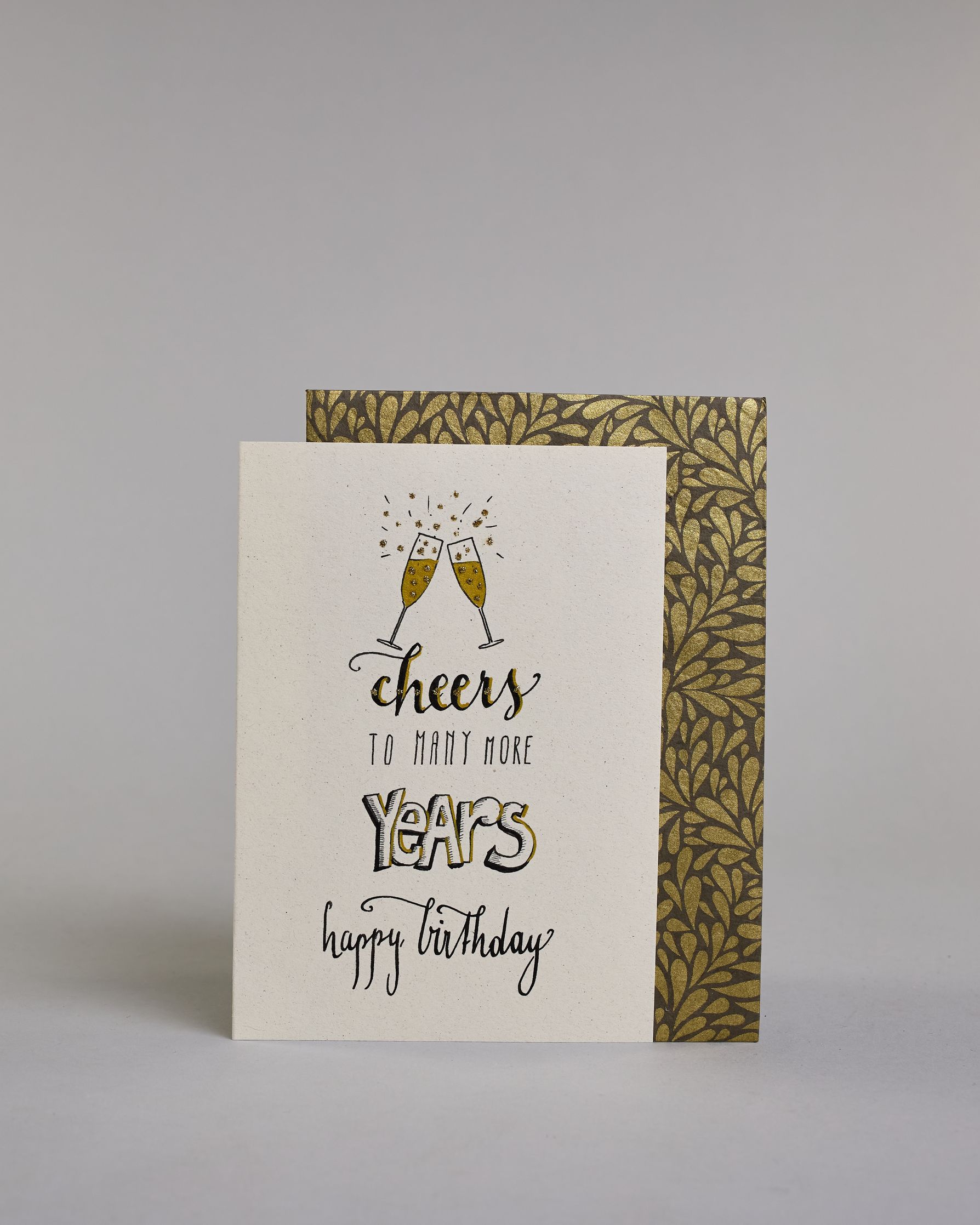 Glückwunschkarte auf Englisch zum Geburtstag mit schwarzer Kalligrafieaufschrift "Cheers to many more years happy birthday", mit gezeichneten miteinander anstoßenden Champagnergläsern über dem Text. Die Zeilen sind wiefolgt aufgeteilt: Unter den Gläsern stehet "cheers" mit geschwungener Schreibschrift, darunter "TO MANY MORE" in dünner gerader Kalligrafieschrift, darunter "YEARS" in dicken weiß gefüllten Blockbuchstaben, darunter "happy birthday" in dünner, kleiner Schreibschrift auf hellem Hintergrund. Da…
