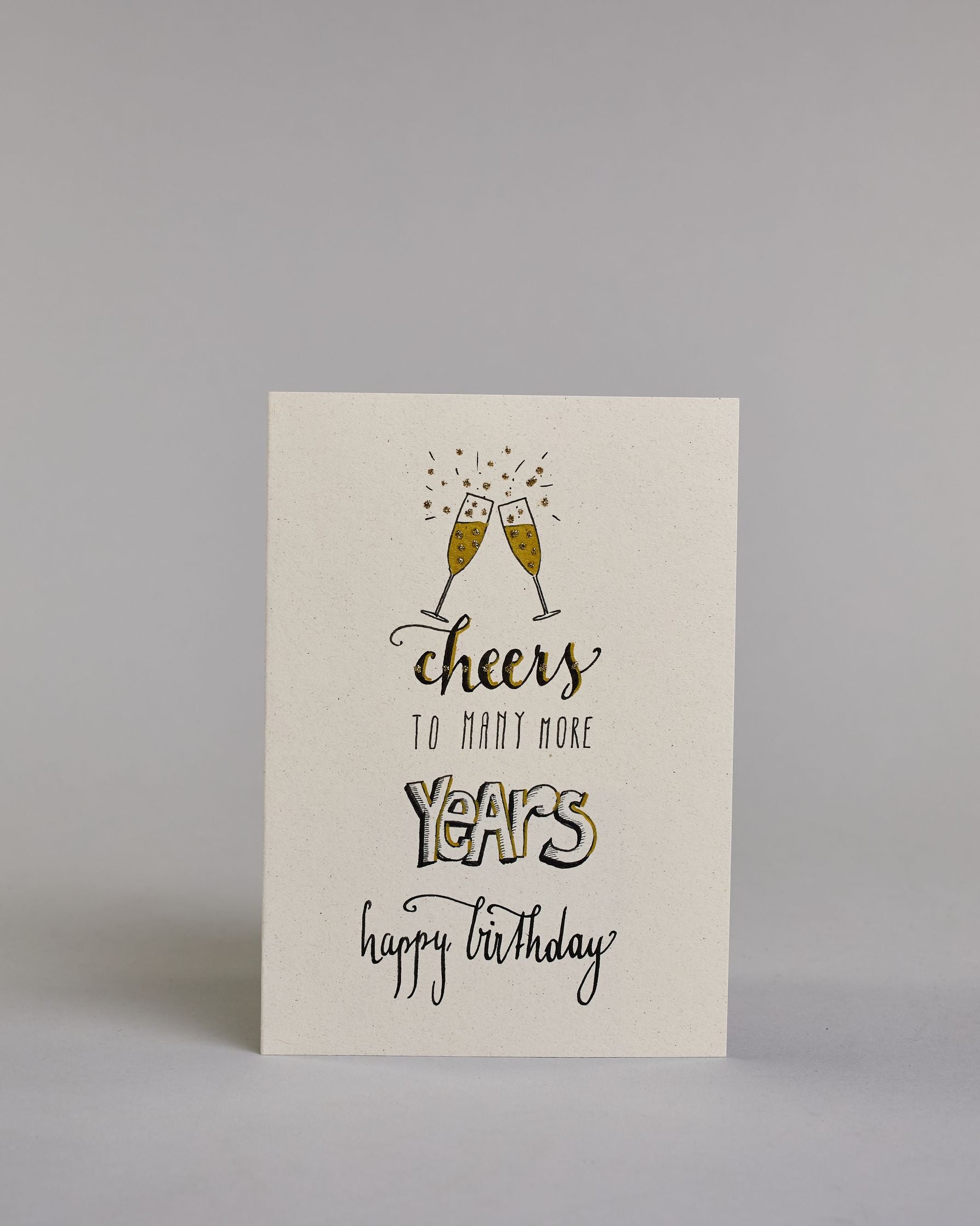 Glückwunschkarte auf Englisch zum Geburtstag mit schwarzer Kalligrafieaufschrift "Cheers to many more years happy birthday", mit gezeichneten miteinander anstoßenden Champagnergläsern über dem Text. Die Zeilen sind wiefolgt aufgeteilt: Unter den Gläsern stehet "cheers" mit geschwungener Schreibschrift, darunter "TO MANY MORE" in dünner gerader Kalligrafieschrift, darunter "YEARS" in dicken weiß gefüllten Blockbuchstaben, darunter "happy birthday" in dünner, kleiner Schreibschrift auf hellem Hintergrund. Da…