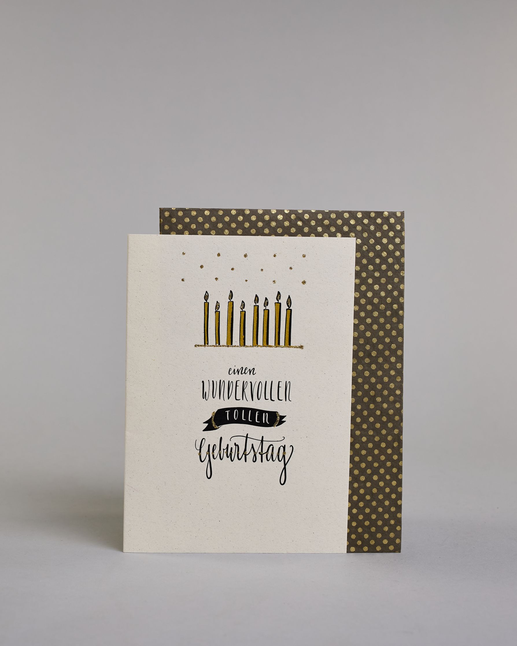 Glückwunschkarte zum Geburtstag mit schwarzer Kalligrafieaufschrift "Einen wundervollen tollen Geburtstag" mit Kerzen über dem Text auf hellem Hintergrund. Die auf vier Zeilen aufgeteilt mit immer jeweils einem Wort. Über den Kerzen sind goldglitzernde Funken abgebildet und das Wort "tollen" befindet sich in einem gezeichnetetn Banner. Dazu ein grün-goldenes handgeschöpftes Kuvert. #2