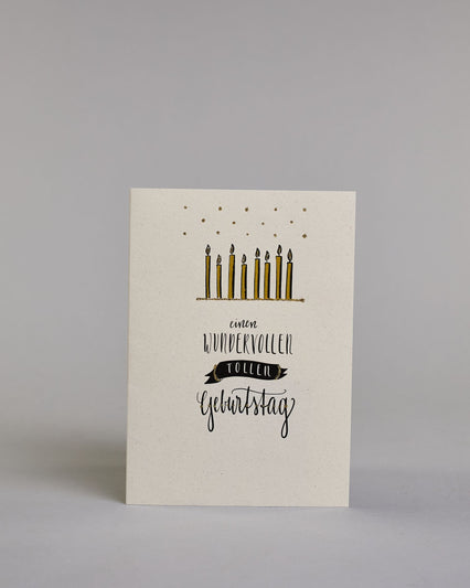 Glückwunschkarte zum Geburtstag mit schwarzer Kalligrafieaufschrift "Einen wundervollen tollen Geburtstag" mit Kerzen über dem Text auf hellem Hintergrund. Die auf vier Zeilen aufgeteilt mit immer jeweils einem Wort. Über den Kerzen sind goldglitzernde Funken abgebildet und das Wort "tollen" befindet sich in einem gezeichnetetn Banner. Dazu ein grün-goldenes handgeschöpftes Kuvert. #1