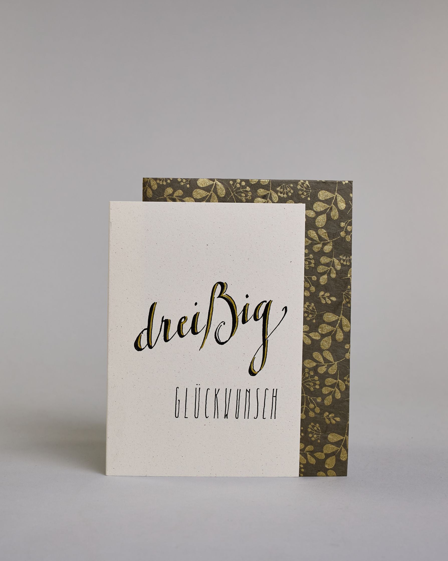 Glückwunschkarte zum Geburtstag mit schwarzer Kalligrafieaufschrift "dreißig Glückwunsch" auf hellem Hintergrund. Die ausgeschriebene Zahl ist leicht aufsteigend in geschwungener, golzverzierter Schreibschrift gestaltet und das Wort "GLÜCKWUNSCH" darunter leicht nach rechts versetzte ist in dünner, gerader Kalligrafieschrift geschrieben. Dazu ein grün-goldenes handgeschöpftes Kuvert. #2