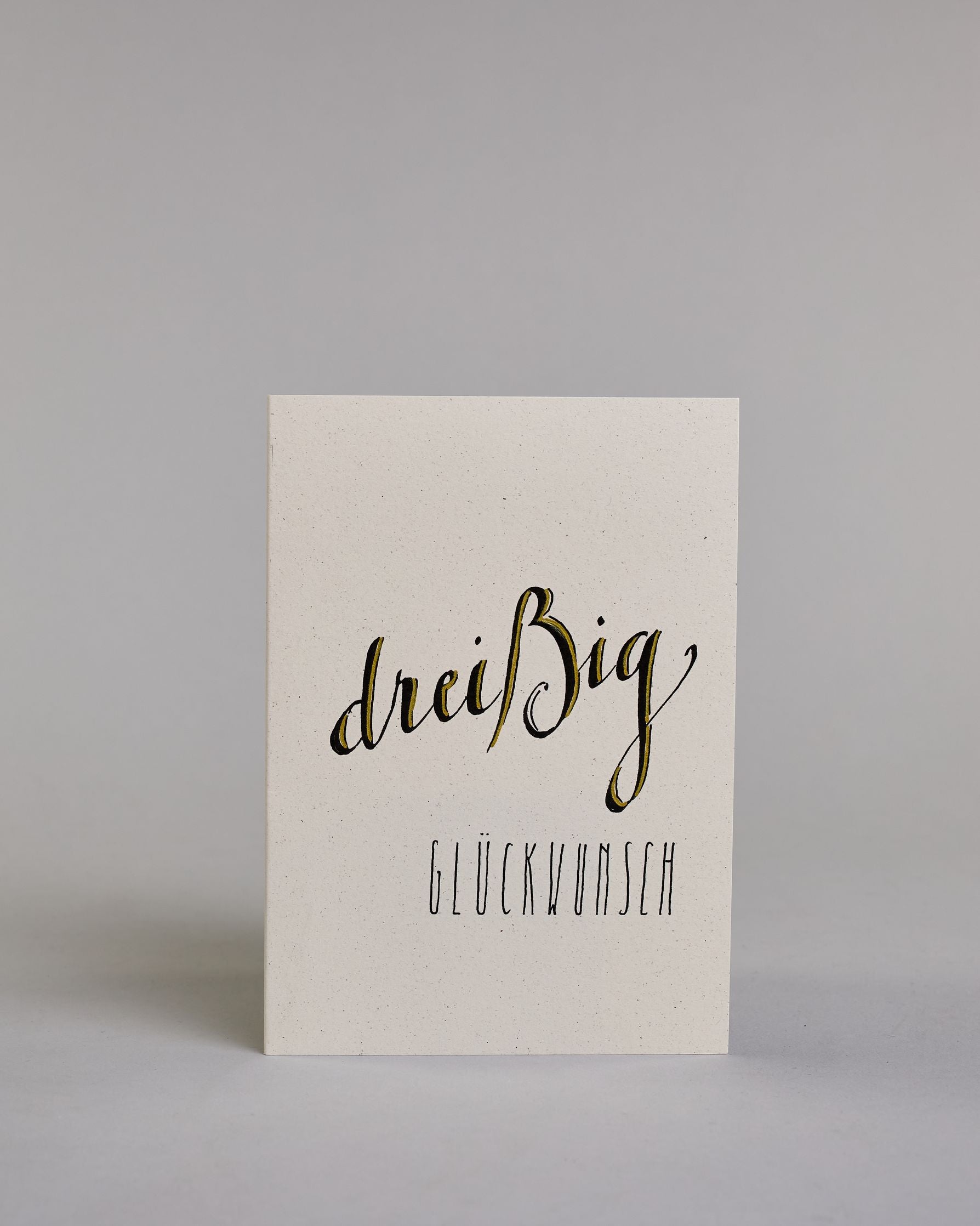 Glückwunschkarte zum Geburtstag mit schwarzer Kalligrafieaufschrift "dreißig Glückwunsch" auf hellem Hintergrund. Die ausgeschriebene Zahl ist leicht aufsteigend in geschwungener, golzverzierter Schreibschrift gestaltet und das Wort "GLÜCKWUNSCH" darunter leicht nach rechts versetzte ist in dünner, gerader Kalligrafieschrift geschrieben. Dazu ein grün-goldenes handgeschöpftes Kuvert. #1