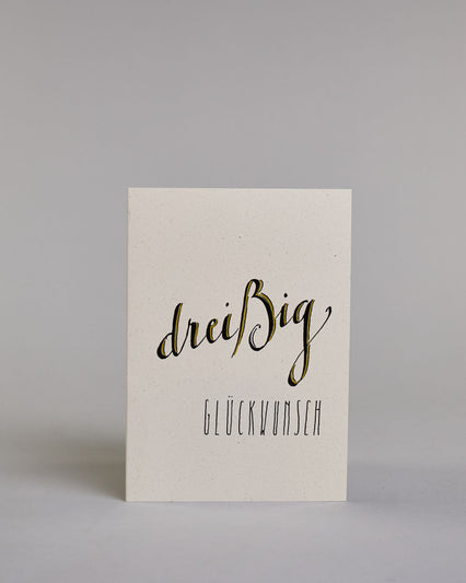 Glückwunschkarte zum Geburtstag mit schwarzer Kalligrafieaufschrift "dreißig Glückwunsch" auf hellem Hintergrund. Die ausgeschriebene Zahl ist leicht aufsteigend in geschwungener, golzverzierter Schreibschrift gestaltet und das Wort "GLÜCKWUNSCH" darunter leicht nach rechts versetzte ist in dünner, gerader Kalligrafieschrift geschrieben. Dazu ein grün-goldenes handgeschöpftes Kuvert. #1