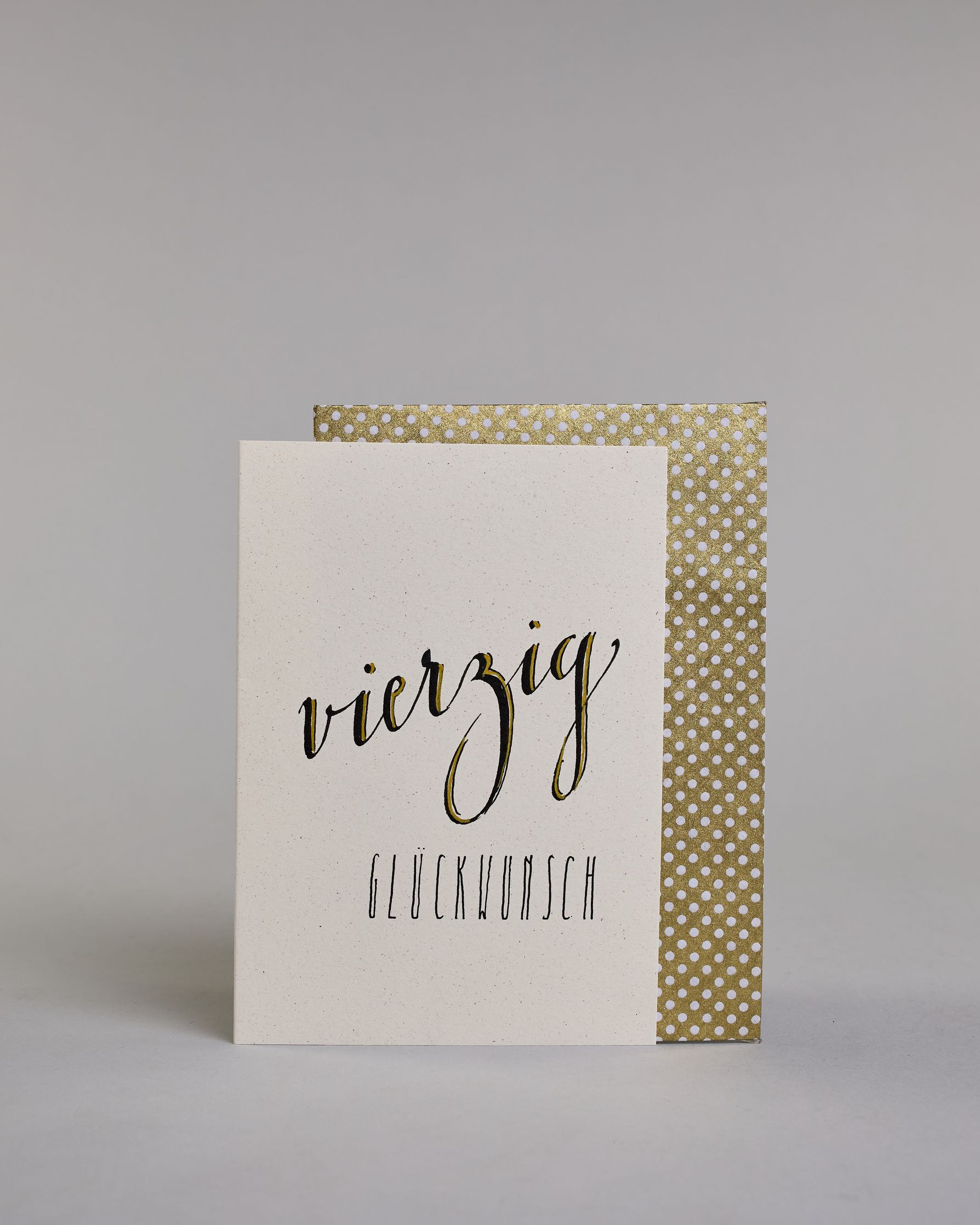 Glückwunschkarte zum Geburtstag mit schwarzer Kalligrafieaufschrift "vierzig Glückwunsch" auf hellem Hintergrund. Die ausgeschriebene Zahl ist leicht aufsteigend in geschwungener, goldverzierter Schreibschrift gestaltet und das Wort "GLÜCKWUNSCH" darunter leicht nach rechts versetzte ist in dünner, gerader Kalligrafieschrift geschrieben. Dazu ein gold-weißes handgeschöpftes Kuvert. #2