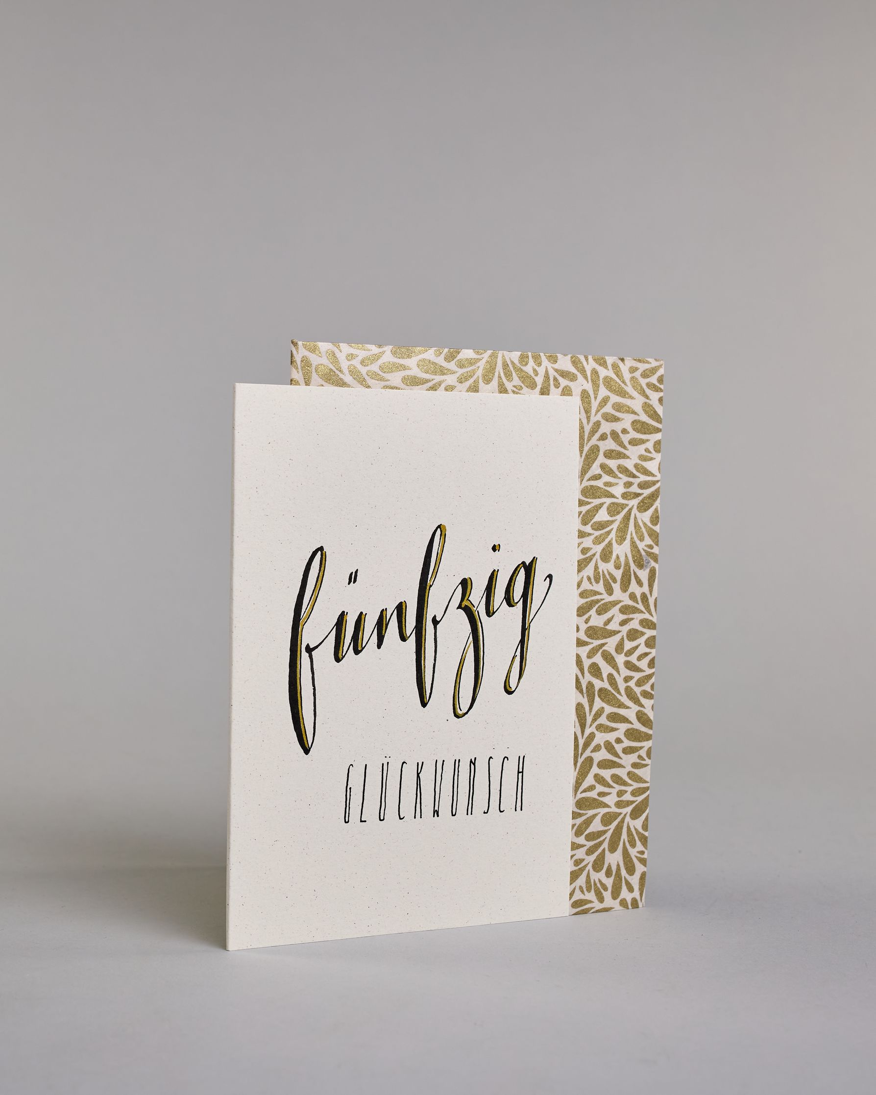 Glückwunschkarte zum Geburtstag mit schwarzer Kalligrafieaufschrift "fünfzig Glückwunsch" auf hellem Hintergrund. Die ausgeschriebene Zahl ist leicht aufsteigend in geschwungener, goldverzierter Schreibschrift gestaltet und das Wort "GLÜCKWUNSCH" darunter leicht nach rechts versetzte ist in dünner, gerader Kalligrafieschrift geschrieben. Dazu ein weiß-goldenes handgeschöpftes Kuvert. #3