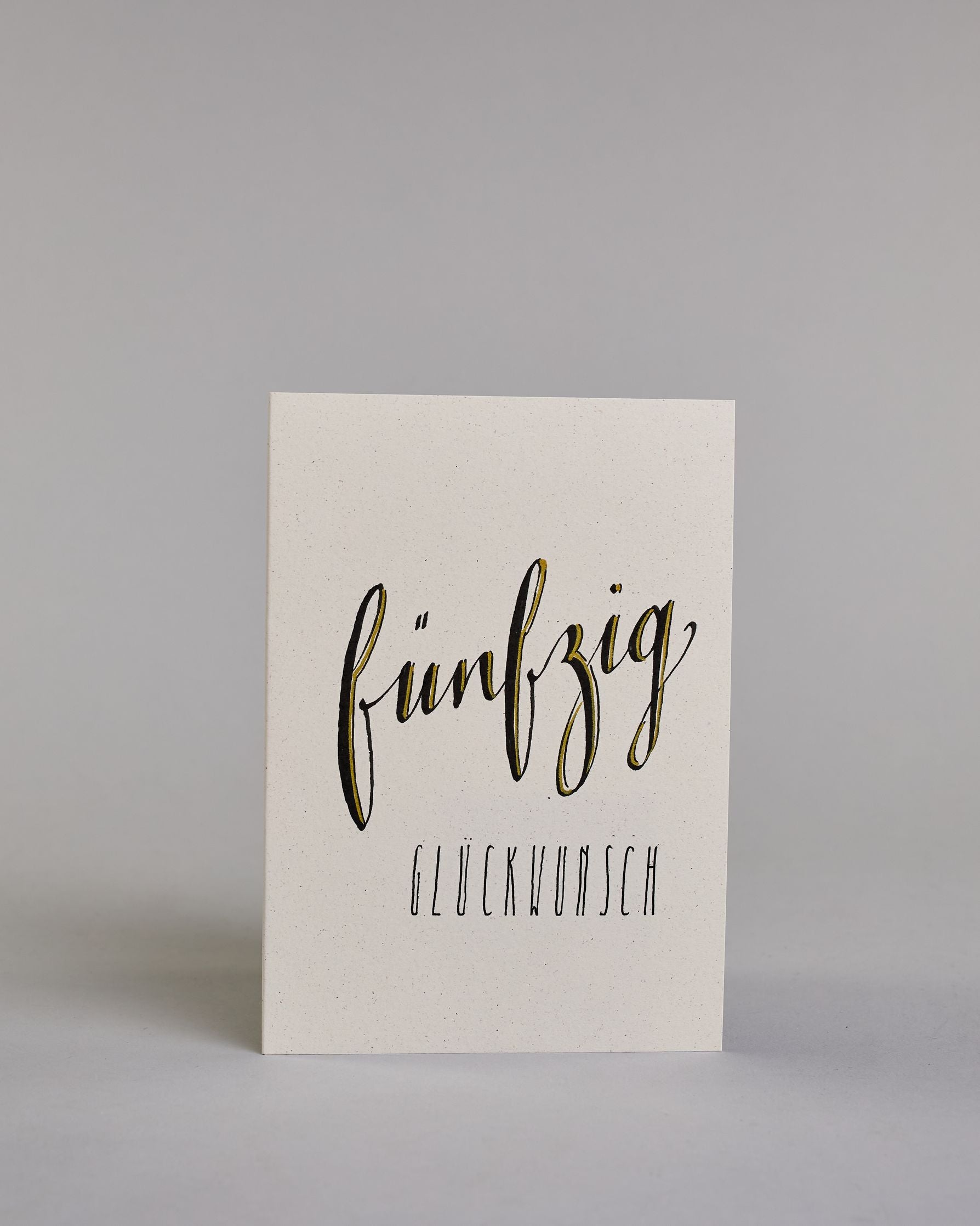 Glückwunschkarte zum Geburtstag mit schwarzer Kalligrafieaufschrift "fünfzig Glückwunsch" auf hellem Hintergrund. Die ausgeschriebene Zahl ist leicht aufsteigend in geschwungener, goldverzierter Schreibschrift gestaltet und das Wort "GLÜCKWUNSCH" darunter leicht nach rechts versetzte ist in dünner, gerader Kalligrafieschrift geschrieben. Dazu ein weiß-goldenes handgeschöpftes Kuvert. #1