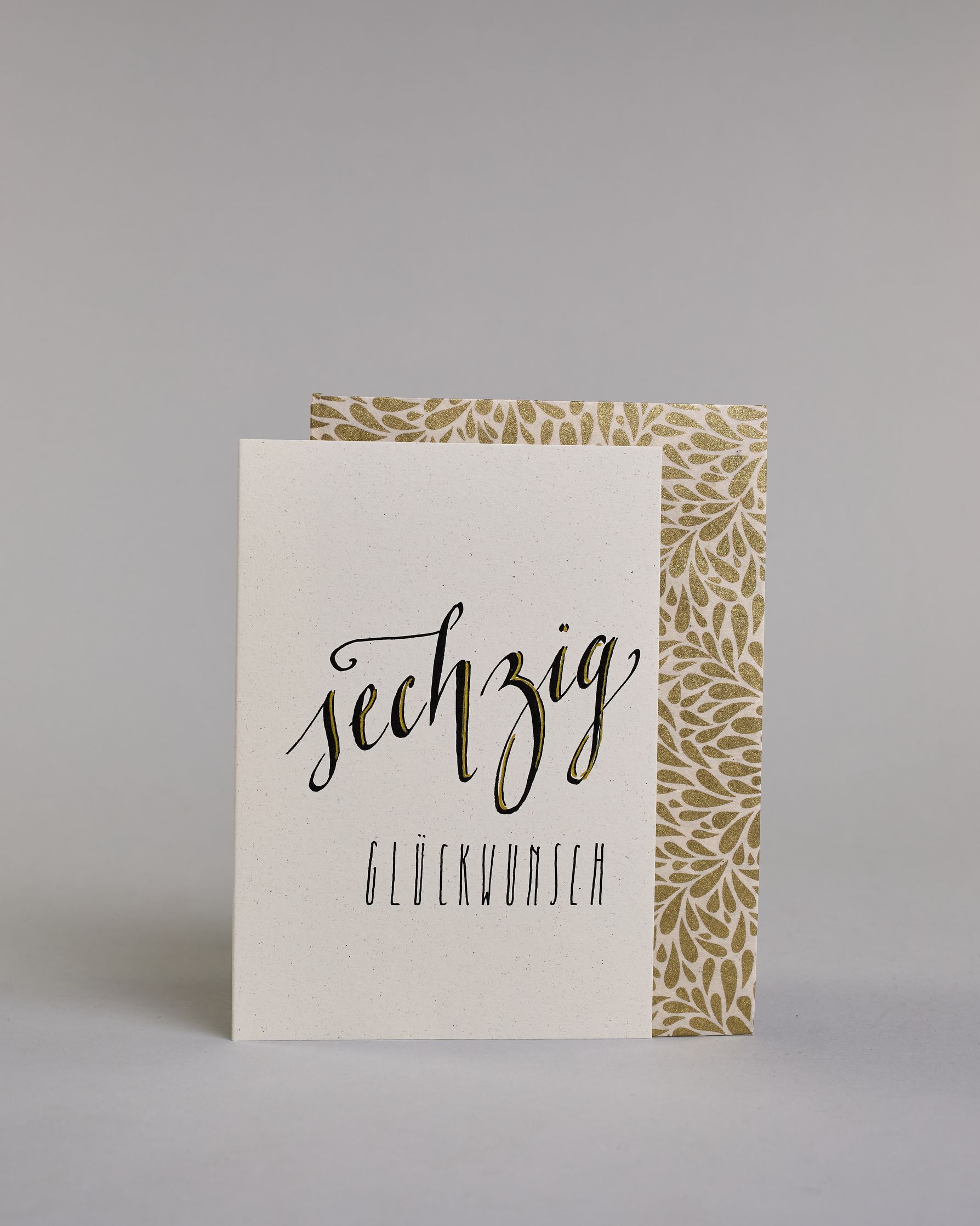 Glückwunschkarte zum Geburtstag mit schwarzer Kalligrafieaufschrift "sechzig Glückwunsch" auf hellem Hintergrund. Die ausgeschriebene Zahl ist leicht aufsteigend in geschwungener, goldverzierter Schreibschrift gestaltet und das Wort "GLÜCKWUNSCH" darunter leicht nach rechts versetzte ist in dünner, gerader Kalligrafieschrift geschrieben. Dazu ein weiß-goldenes handgeschöpftes Kuvert. #2