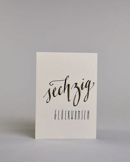 Glückwunschkarte zum Geburtstag mit schwarzer Kalligrafieaufschrift "sechzig Glückwunsch" auf hellem Hintergrund. Die ausgeschriebene Zahl ist leicht aufsteigend in geschwungener, goldverzierter Schreibschrift gestaltet und das Wort "GLÜCKWUNSCH" darunter leicht nach rechts versetzte ist in dünner, gerader Kalligrafieschrift geschrieben. Dazu ein weiß-goldenes handgeschöpftes Kuvert. #1