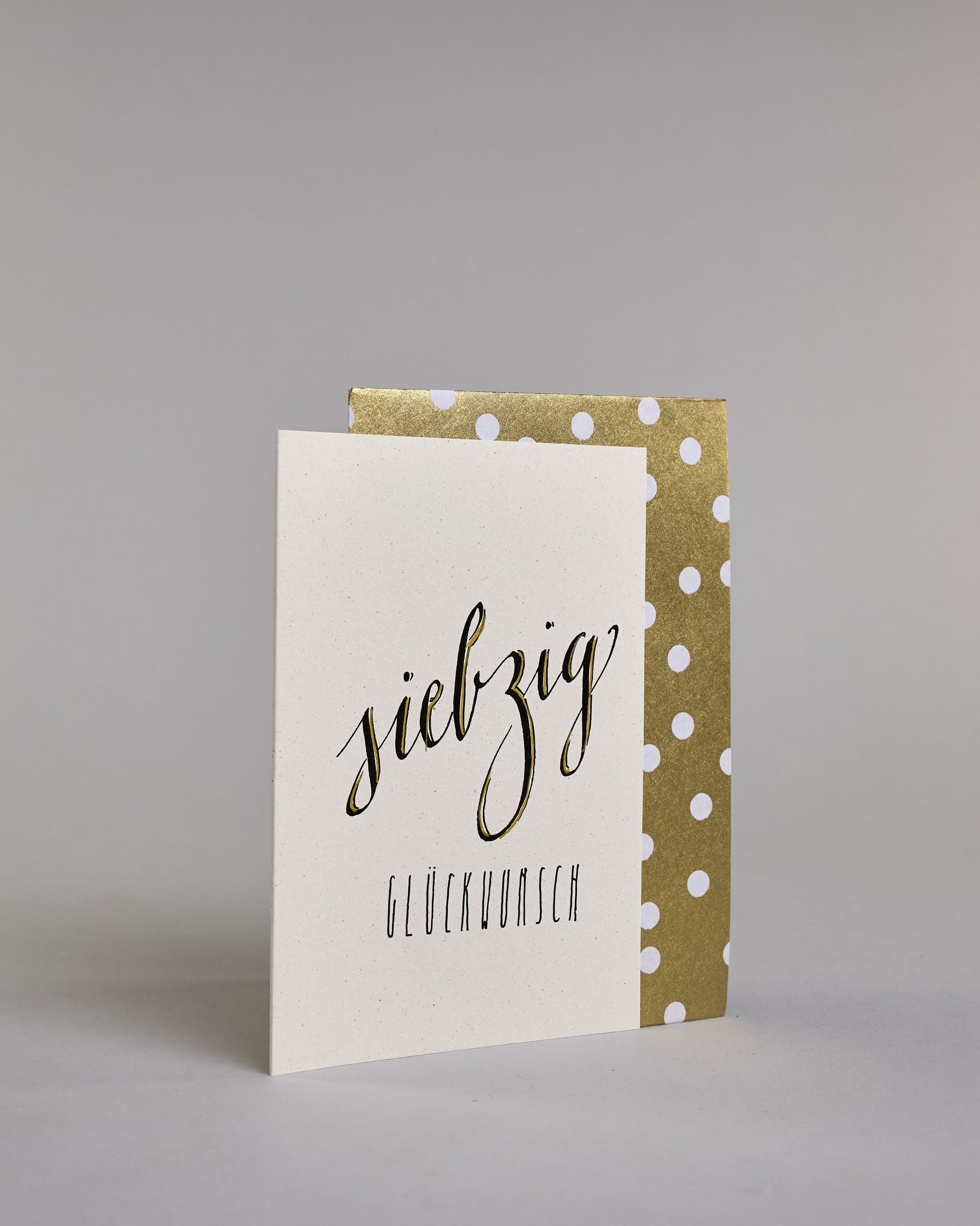 Glückwunschkarte zum Geburtstag mit schwarzer Kalligrafieaufschrift "siebzig Glückwunsch" auf hellem Hintergrund. Die ausgeschriebene Zahl ist leicht aufsteigend in geschwungener, goldverzierter Schreibschrift gestaltet und das Wort "GLÜCKWUNSCH" darunter leicht nach rechts versetzte ist in dünner, gerader Kalligrafieschrift geschrieben. Dazu ein gold-weißes handgeschöpftes Kuvert. #3