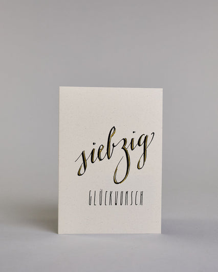 Glückwunschkarte zum Geburtstag mit schwarzer Kalligrafieaufschrift "siebzig Glückwunsch" auf hellem Hintergrund. Die ausgeschriebene Zahl ist leicht aufsteigend in geschwungener, goldverzierter Schreibschrift gestaltet und das Wort "GLÜCKWUNSCH" darunter leicht nach rechts versetzte ist in dünner, gerader Kalligrafieschrift geschrieben. Dazu ein gold-weißes handgeschöpftes Kuvert. #1