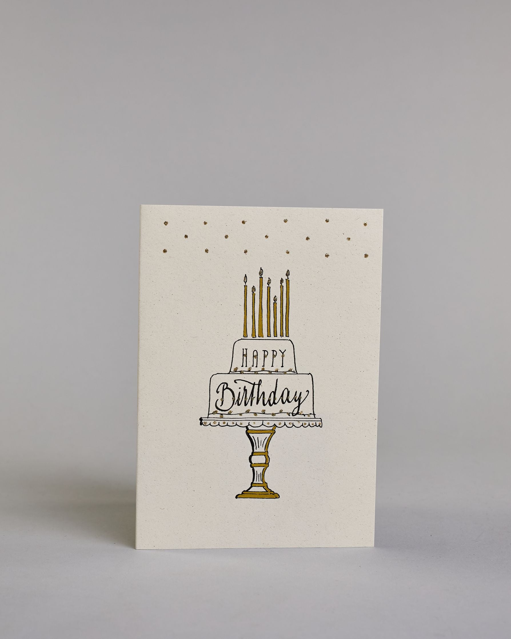 Glückwunschkarte auf Englisch zum Geburtstag mit schwarzer Kalligrafieaufschrift "Happy Birthday" in einer gezeichneten zweistöckigen Geburtstagstorte auf einem Tortenständer mit sieben Kerzen auf der Torte auf hellem Hintergrund. Das Wort "HAPPY" ist in dünner geraden Kalligrafieschrift in der oberen Stock der Torte geschrieben, das "Birthday" in den unteren Stock mit geschwungener Schreibschrift. Sowohl Kuchen, Tortenstände, Kerzen und Stift sind mit gold und goldglitzer verziert. Dazu ein grün-goldenes…