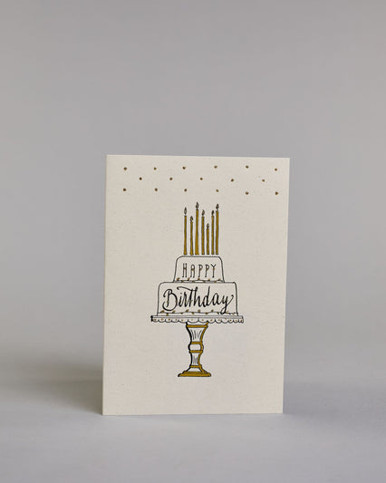 Glückwunschkarte auf Englisch zum Geburtstag mit schwarzer Kalligrafieaufschrift "Happy Birthday" in einer gezeichneten zweistöckigen Geburtstagstorte auf einem Tortenständer mit sieben Kerzen auf der Torte auf hellem Hintergrund. Das Wort "HAPPY" ist in dünner geraden Kalligrafieschrift in der oberen Stock der Torte geschrieben, das "Birthday" in den unteren Stock mit geschwungener Schreibschrift. Sowohl Kuchen, Tortenstände, Kerzen und Stift sind mit gold und goldglitzer verziert. Dazu ein grün-goldenes…