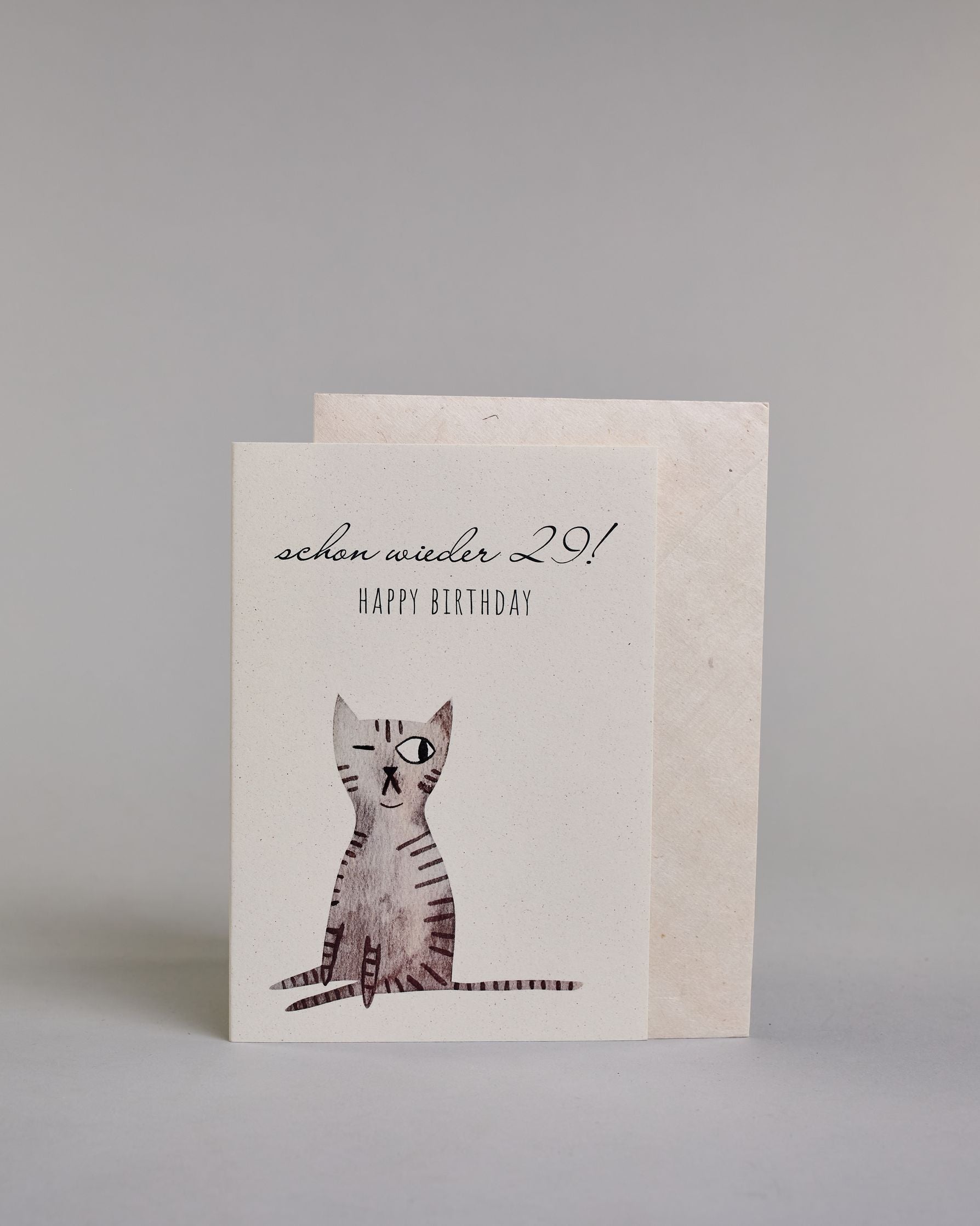 Glückwunschkarte zum Geburtstag mit schwarzer Kalligrafieaufschrift "Schon wieder 29! Happy Birthday", darunter eine gezeichnete grau, schwarz getigerte Katze die mit dem linken Auge zwinkert auf hellem Hintergrund. Der Text ist auf zwei Zeilen aufgeteilt. In der ersten steht "schon wieder 29!" in einer geschwungenen Kalligrafieschrift und darunter "HAPPY BIRTHDAY" in dünner gerader Schrift. Dazu ein weißes handgeschöpftes Kuvert. #2