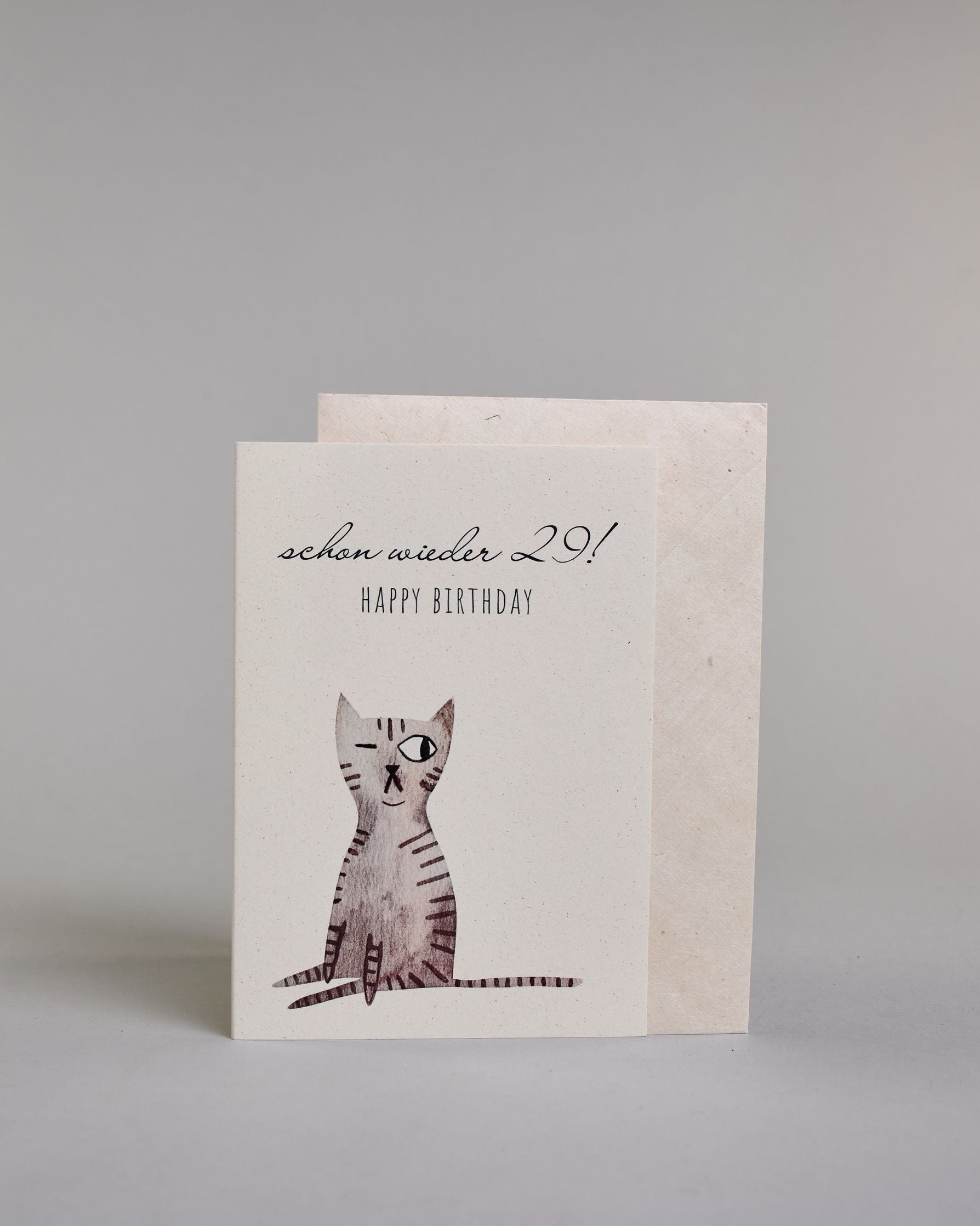 Glückwunschkarte zum Geburtstag mit schwarzer Kalligrafieaufschrift "Schon wieder 29! Happy Birthday", darunter eine gezeichnete grau, schwarz getigerte Katze die mit dem linken Auge zwinkert auf hellem Hintergrund. Der Text ist auf zwei Zeilen aufgeteilt. In der ersten steht "schon wieder 29!" in einer geschwungenen Kalligrafieschrift und darunter "HAPPY BIRTHDAY" in dünner gerader Schrift. Dazu ein weißes handgeschöpftes Kuvert. #3