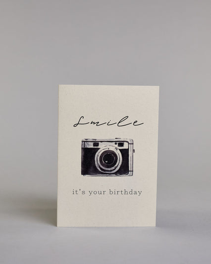 Glückwunschkarte auf Englisch zum Geburtstag mit schwarzer Kalligrafieaufschrift "Smile it's your birthday", mit einer gezeichneten schwarz-silbernen Kamera auf hellem Hintergrund. Die Kamera ist mittig auf der Karte, darüber steht in geschwungener Kalligrafieschrift "smile" und unter der Kamera in gerader Schreibmaschinenschrift "it's your birthday". Dazu ein weißes handgeschöpftes Kuvert. #1