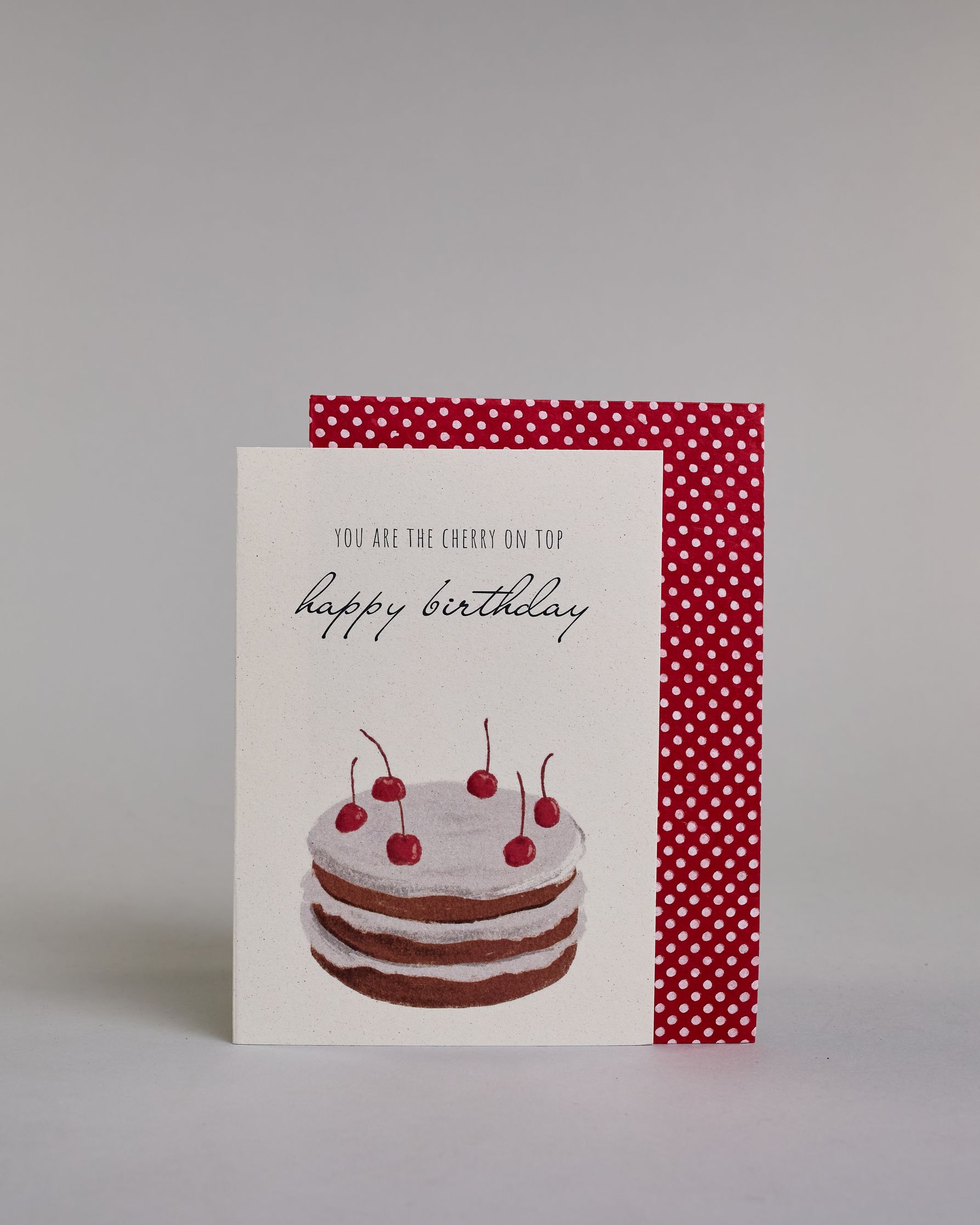 Glückwunschkarte auf Englisch zum Geburtstag mit schwarzer Kalligrafieaufschrift "you are the cherry on top happy birthday", mit einer mit gezeichneten mit sechs Kirschen besetzten Torte auf hellem Hintergrund. Der Text ist auf zwei Zeilen aufgeteilt. Oben steht in dünner gerader Kalligrafieschrift "YOU ARE THE CHERRY ON TOP", darunter "happy birthday" in geschwungener Handschrift und darunter ist der Kuchen der aus drei braun gebackenen Schicken besteht, mit jeweils einer weißen Cremeschicht dazwischen un…