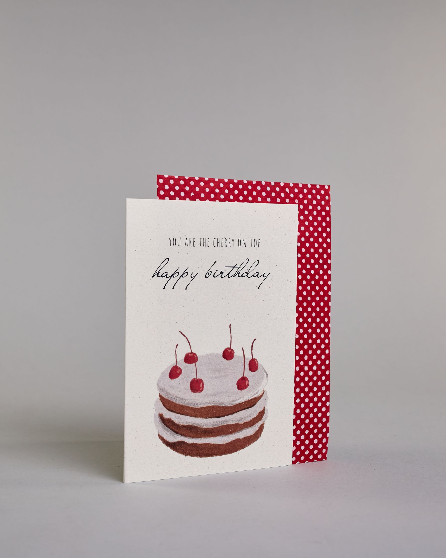 Glückwunschkarte auf Englisch zum Geburtstag mit schwarzer Kalligrafieaufschrift "you are the cherry on top happy birthday", mit einer mit gezeichneten mit sechs Kirschen besetzten Torte auf hellem Hintergrund. Der Text ist auf zwei Zeilen aufgeteilt. Oben steht in dünner gerader Kalligrafieschrift "YOU ARE THE CHERRY ON TOP", darunter "happy birthday" in geschwungener Handschrift und darunter ist der Kuchen der aus drei braun gebackenen Schicken besteht, mit jeweils einer weißen Cremeschicht dazwischen un…