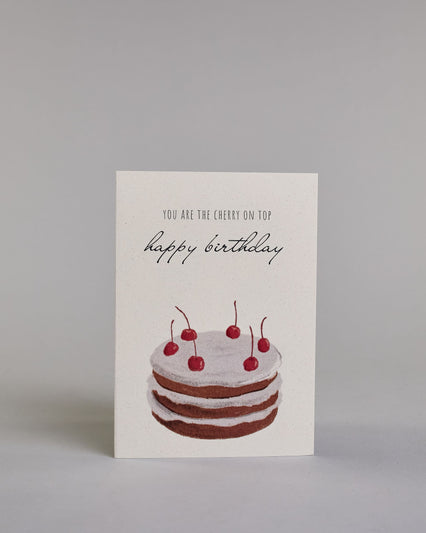 Glückwunschkarte auf Englisch zum Geburtstag mit schwarzer Kalligrafieaufschrift "you are the cherry on top happy birthday", mit einer mit gezeichneten mit sechs Kirschen besetzten Torte auf hellem Hintergrund. Der Text ist auf zwei Zeilen aufgeteilt. Oben steht in dünner gerader Kalligrafieschrift "YOU ARE THE CHERRY ON TOP", darunter "happy birthday" in geschwungener Handschrift und darunter ist der Kuchen der aus drei braun gebackenen Schicken besteht, mit jeweils einer weißen Cremeschicht dazwischen un…