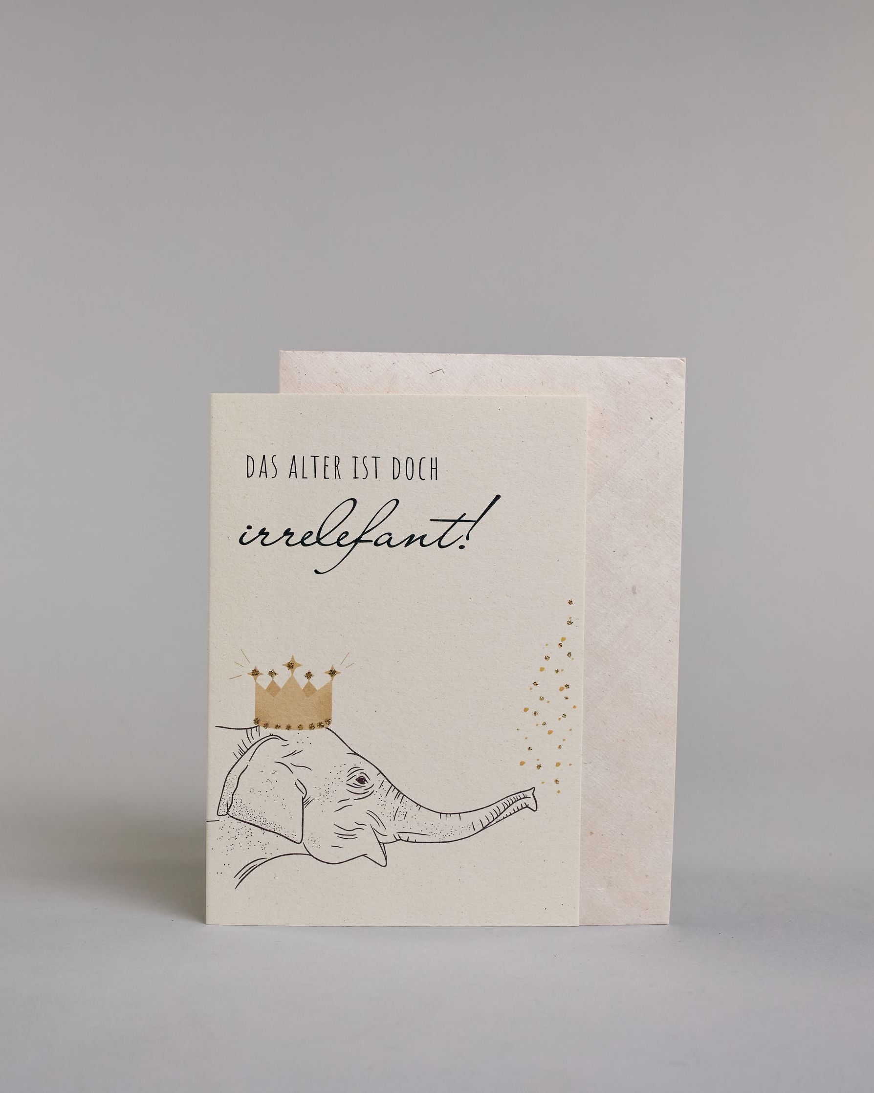 Glückwunschkarte zum Geburtstag mit schwarzer Kalligrafieaufschrift "Das Alter ist doch irrelefant!", mit einem gezeichneten Elefanten mit einer Krone auf dem Kopf, der Kofetti aus dem Rüssel pustet auf hellem Hintergrund. Oben steht in dünner gerader Kalligrafieschrift "DAS ALTER IST DOCH", darunter "irrelefant" in geschwungener Handschrift mit einem "f" statt einem "v" für das Wortspiel. Darunter ist der Elefant dessen Kopf von der linken Seite auf die Karte kommt. Dazu ein grün-goldenes handgeschöpftes…