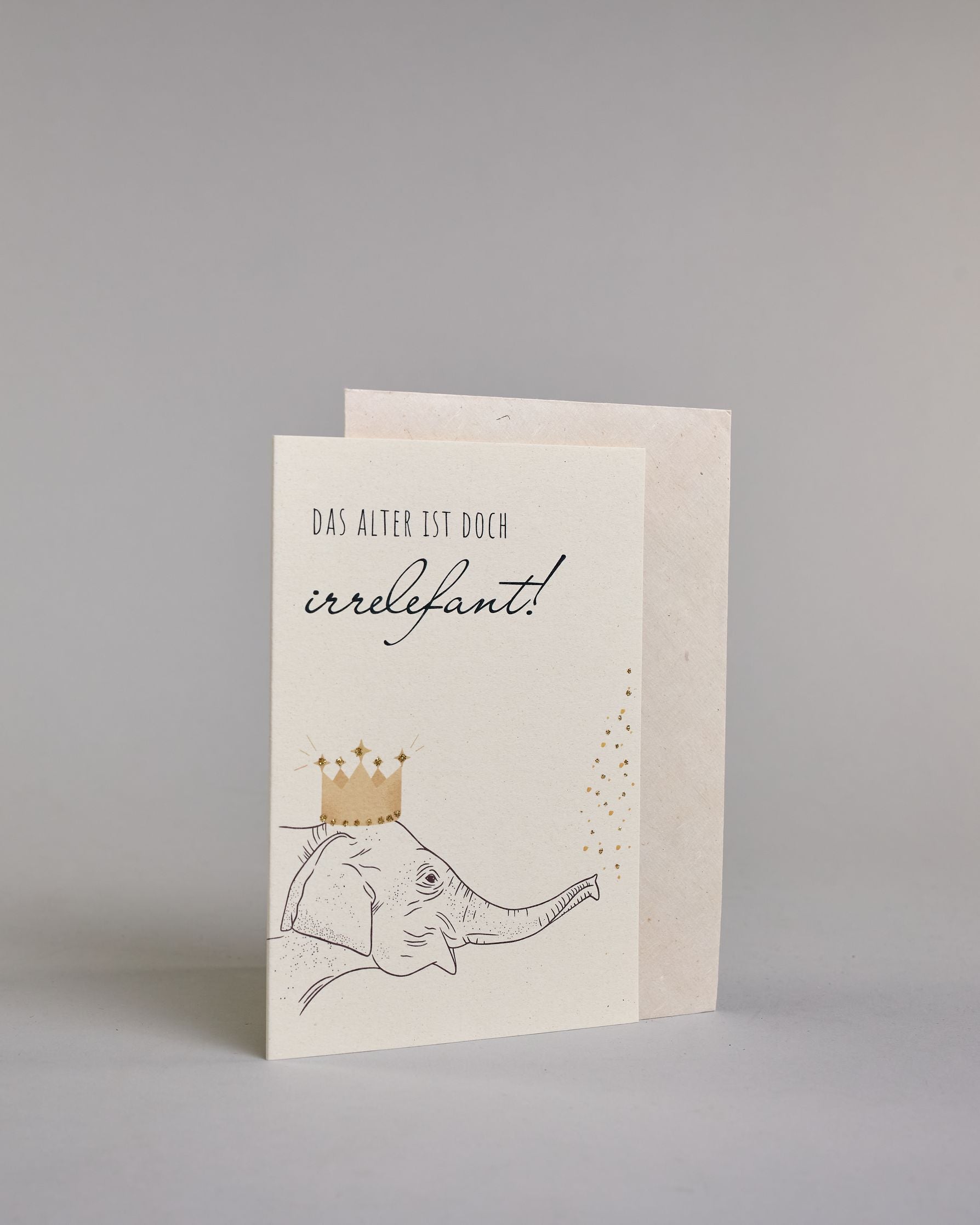 Glückwunschkarte zum Geburtstag mit schwarzer Kalligrafieaufschrift "Das Alter ist doch irrelefant!", mit einem gezeichneten Elefanten mit einer Krone auf dem Kopf, der Kofetti aus dem Rüssel pustet auf hellem Hintergrund. Oben steht in dünner gerader Kalligrafieschrift "DAS ALTER IST DOCH", darunter "irrelefant" in geschwungener Handschrift mit einem "f" statt einem "v" für das Wortspiel. Darunter ist der Elefant dessen Kopf von der linken Seite auf die Karte kommt. Dazu ein grün-goldenes handgeschöpftes…