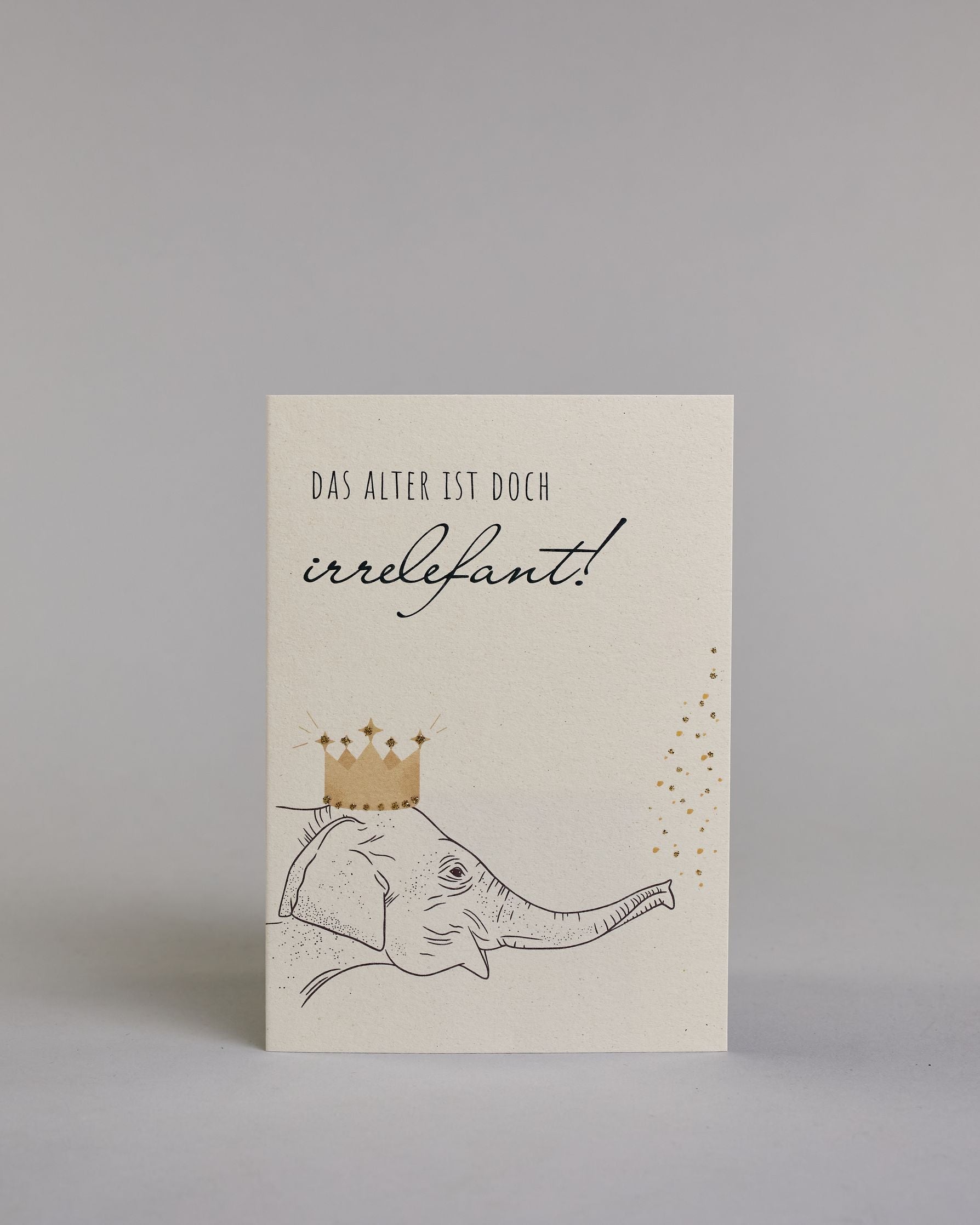 Glückwunschkarte zum Geburtstag mit schwarzer Kalligrafieaufschrift "Das Alter ist doch irrelefant!", mit einem gezeichneten Elefanten mit einer Krone auf dem Kopf, der Kofetti aus dem Rüssel pustet auf hellem Hintergrund. Oben steht in dünner gerader Kalligrafieschrift "DAS ALTER IST DOCH", darunter "irrelefant" in geschwungener Handschrift mit einem "f" statt einem "v" für das Wortspiel. Darunter ist der Elefant dessen Kopf von der linken Seite auf die Karte kommt. Dazu ein grün-goldenes handgeschöpftes…