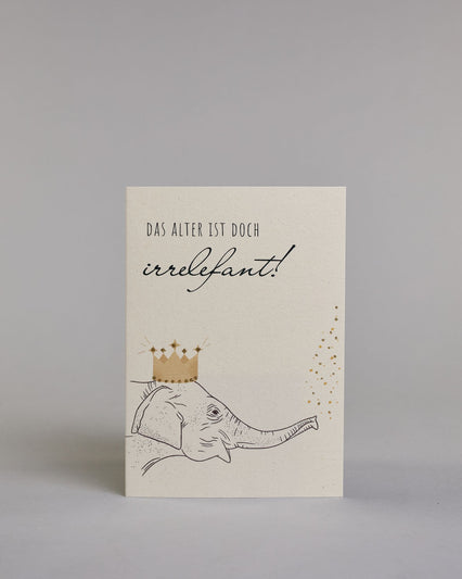 Glückwunschkarte zum Geburtstag mit schwarzer Kalligrafieaufschrift "Das Alter ist doch irrelefant!", mit einem gezeichneten Elefanten mit einer Krone auf dem Kopf, der Kofetti aus dem Rüssel pustet auf hellem Hintergrund. Oben steht in dünner gerader Kalligrafieschrift "DAS ALTER IST DOCH", darunter "irrelefant" in geschwungener Handschrift mit einem "f" statt einem "v" für das Wortspiel. Darunter ist der Elefant dessen Kopf von der linken Seite auf die Karte kommt. Dazu ein grün-goldenes handgeschöpftes…