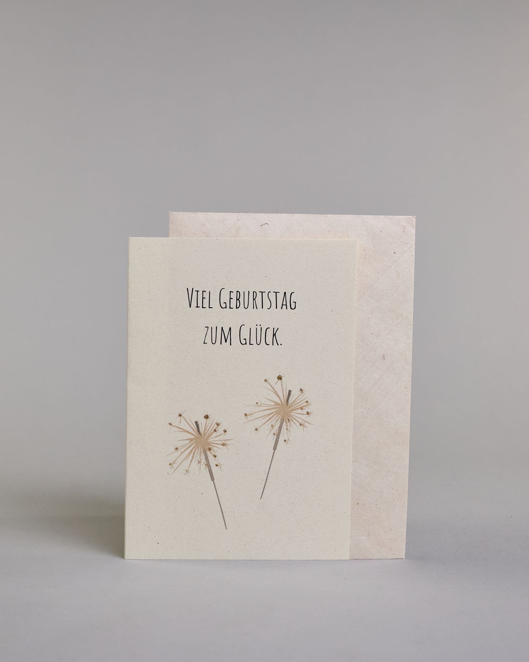 Glückwunschkarte zum Geburtstag mit schwarzer Kalligrafieaufschrift "Viel Geburtstag zum Glück" mit goldenen gezeichneten Sprühkerzen auf hellem Hintergrund. Das Wortspiel ist auf zwei Zeilen aufgeteilt, welche jeweils immer zwei Wort beinhalten, also "VIEL GEBURTSTAG" und darunter "ZUM GLÜCK". Die darunterliegenden Sprühkerzen sind mit goldglitzer verfeinert um die Funken zu verdeutlichen. Die Schrift ist eine dünne gerade Kalligrafieschrift. Dazu ein weißes handgeschöpftes Kuvert. #2