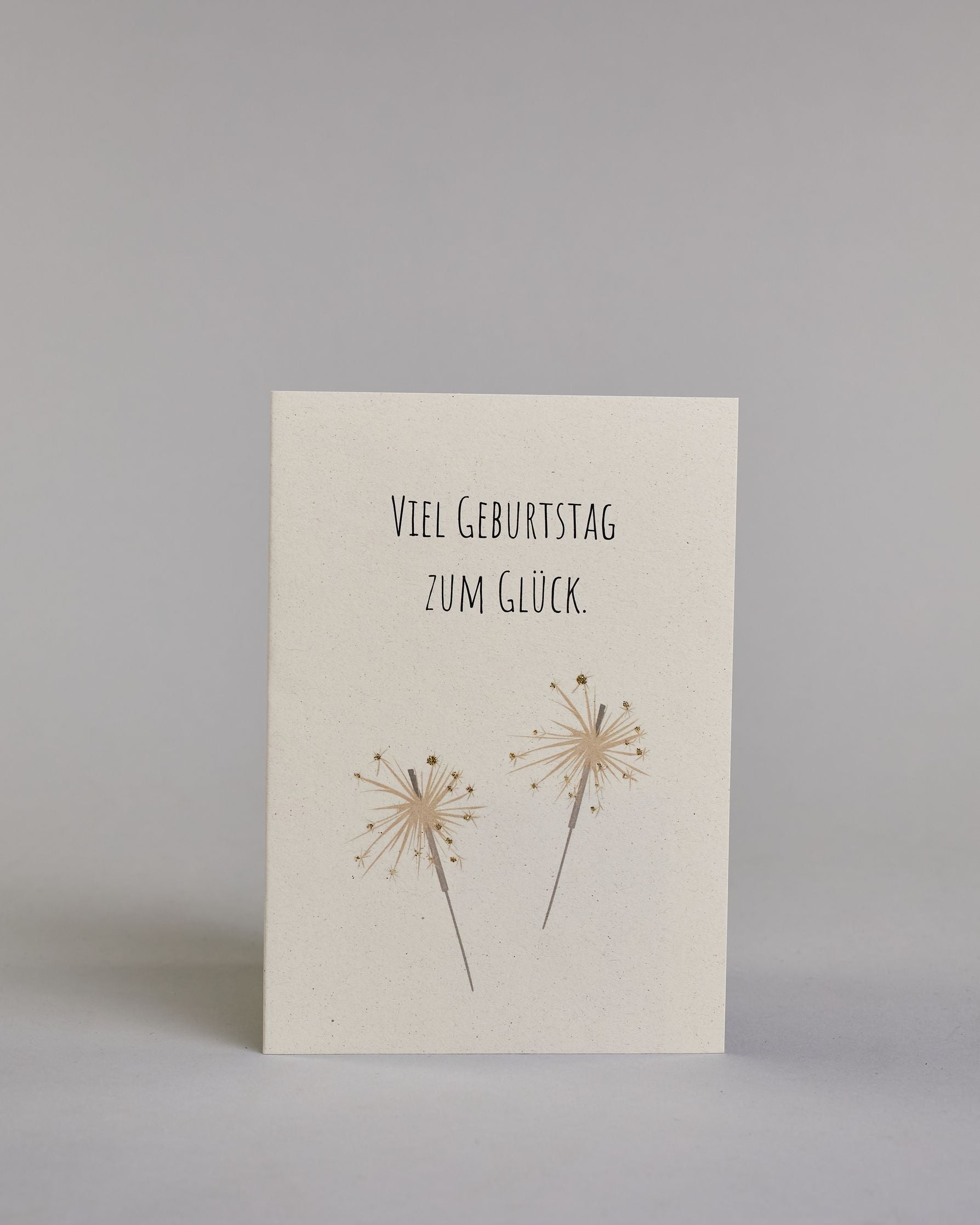 Glückwunschkarte zum Geburtstag mit schwarzer Kalligrafieaufschrift "Viel Geburtstag zum Glück" mit goldenen gezeichneten Sprühkerzen auf hellem Hintergrund. Das Wortspiel ist auf zwei Zeilen aufgeteilt, welche jeweils immer zwei Wort beinhalten, also "VIEL GEBURTSTAG" und darunter "ZUM GLÜCK". Die darunterliegenden Sprühkerzen sind mit goldglitzer verfeinert um die Funken zu verdeutlichen. Die Schrift ist eine dünne gerade Kalligrafieschrift. Dazu ein weißes handgeschöpftes Kuvert. #1