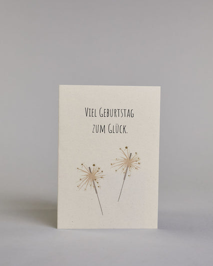 Glückwunschkarte zum Geburtstag mit schwarzer Kalligrafieaufschrift "Viel Geburtstag zum Glück" mit goldenen gezeichneten Sprühkerzen auf hellem Hintergrund. Das Wortspiel ist auf zwei Zeilen aufgeteilt, welche jeweils immer zwei Wort beinhalten, also "VIEL GEBURTSTAG" und darunter "ZUM GLÜCK". Die darunterliegenden Sprühkerzen sind mit goldglitzer verfeinert um die Funken zu verdeutlichen. Die Schrift ist eine dünne gerade Kalligrafieschrift. Dazu ein weißes handgeschöpftes Kuvert. #1